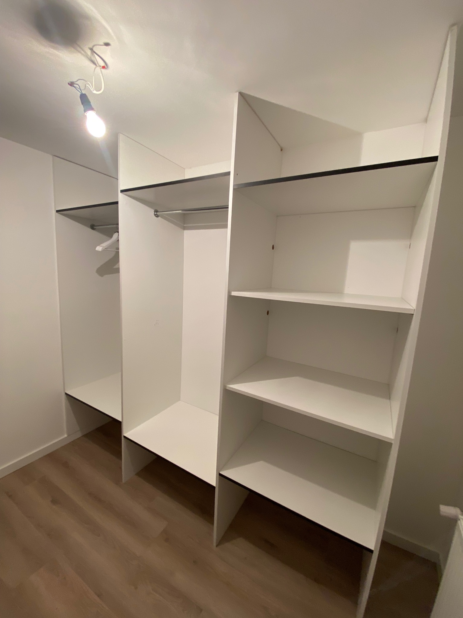 Wnętrze garderoby z białymi półkami i drążkami na ubrania, widoczna żarówka bez klosza, drewniana podłoga.