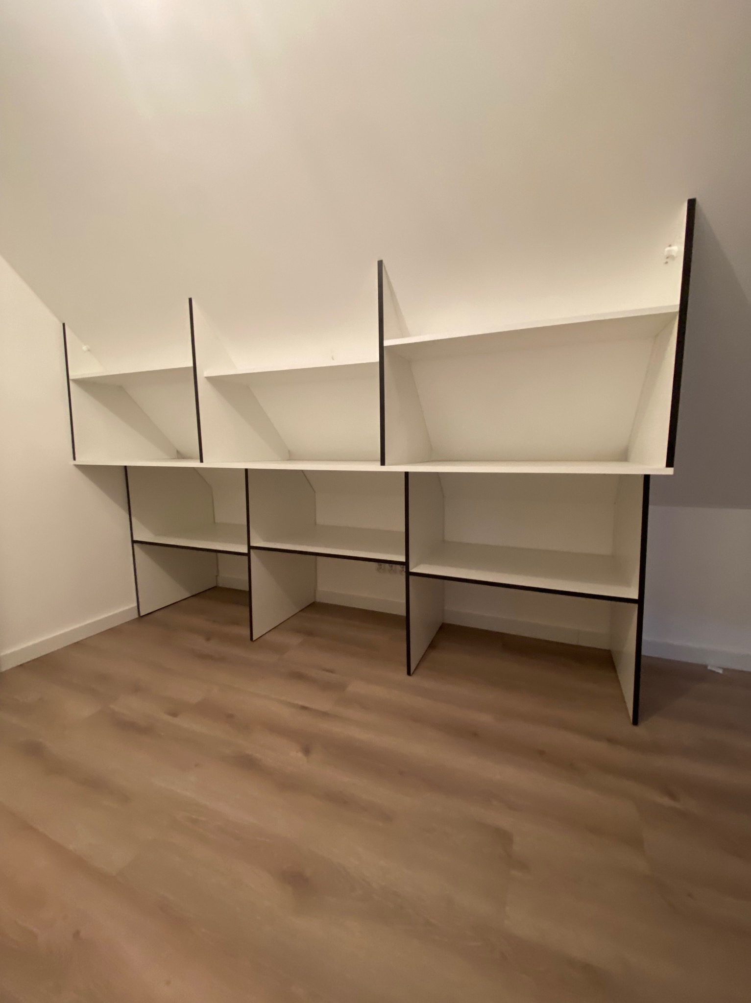 Białe półki z czarnymi obramowaniami, zamontowane pod skosem dachu, w pustym pomieszczeniu z panelami podłogowymi.