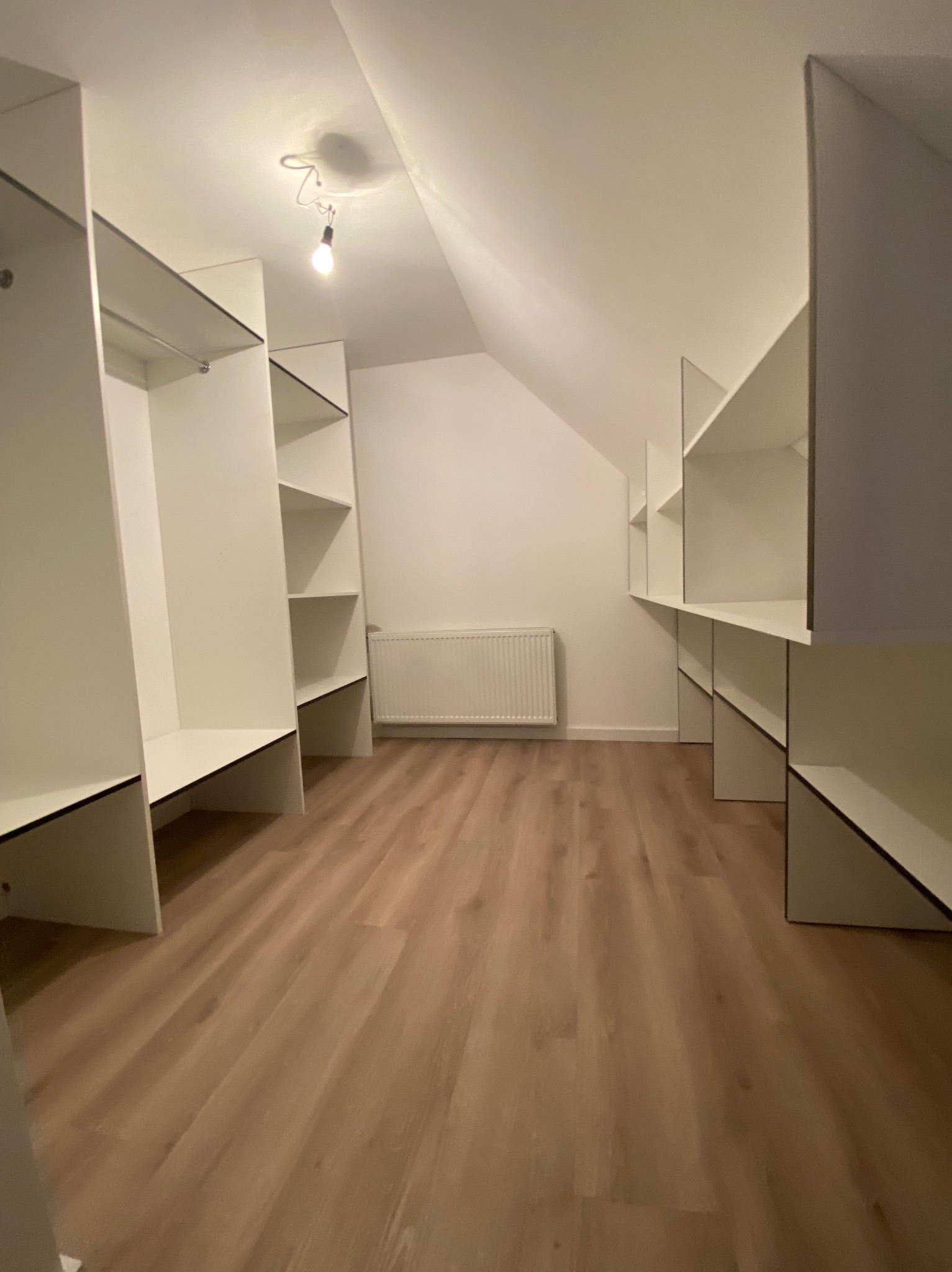 Pusta garderoba w skosach poddasza, białe półki i wieszaki zamontowane na ścianach, widoczny grzejnik i drewniana podłoga.