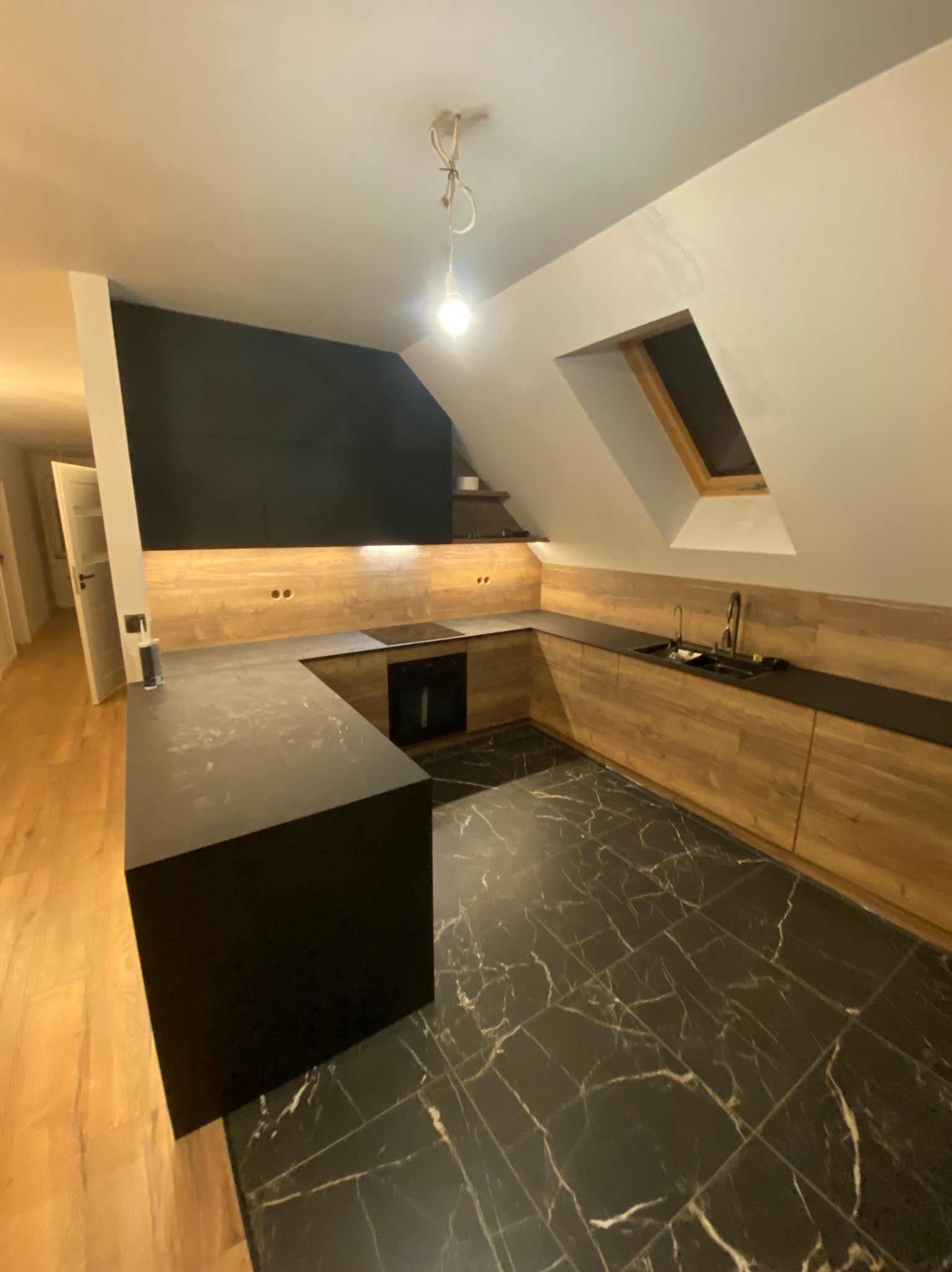 Kuchnia w skosach z czarnymi blatami i drewnopodobnymi frontami, czarna podłoga imitująca marmur, widoczne niedokończone podłączenie lampy.