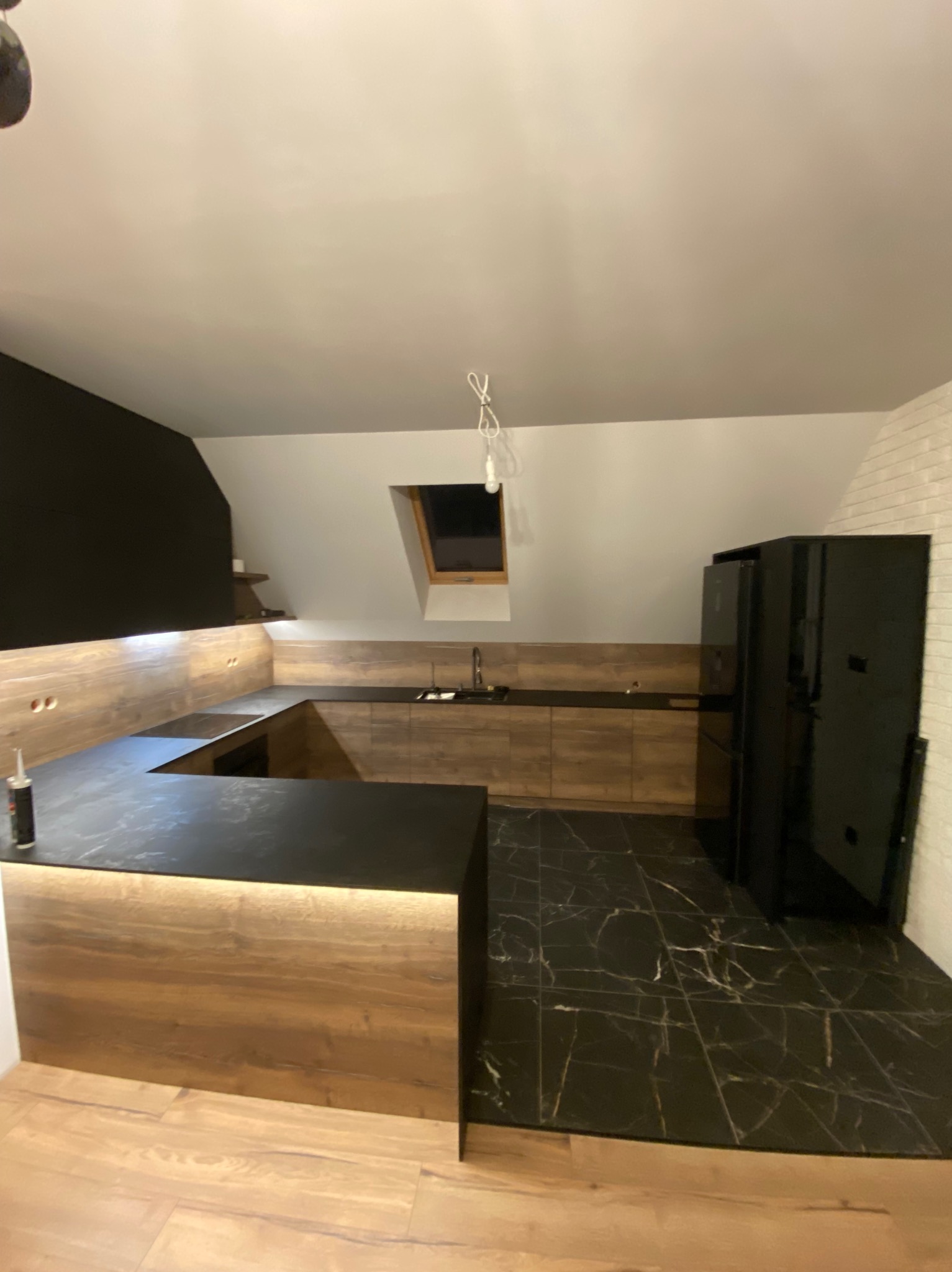 Kuchenna zabudowa w skosach poddasza z czarnym blatem i drewnopodobnymi frontami, czarna podłoga z marmurkowym wzorem, widoczna tuba silikonu na blacie.