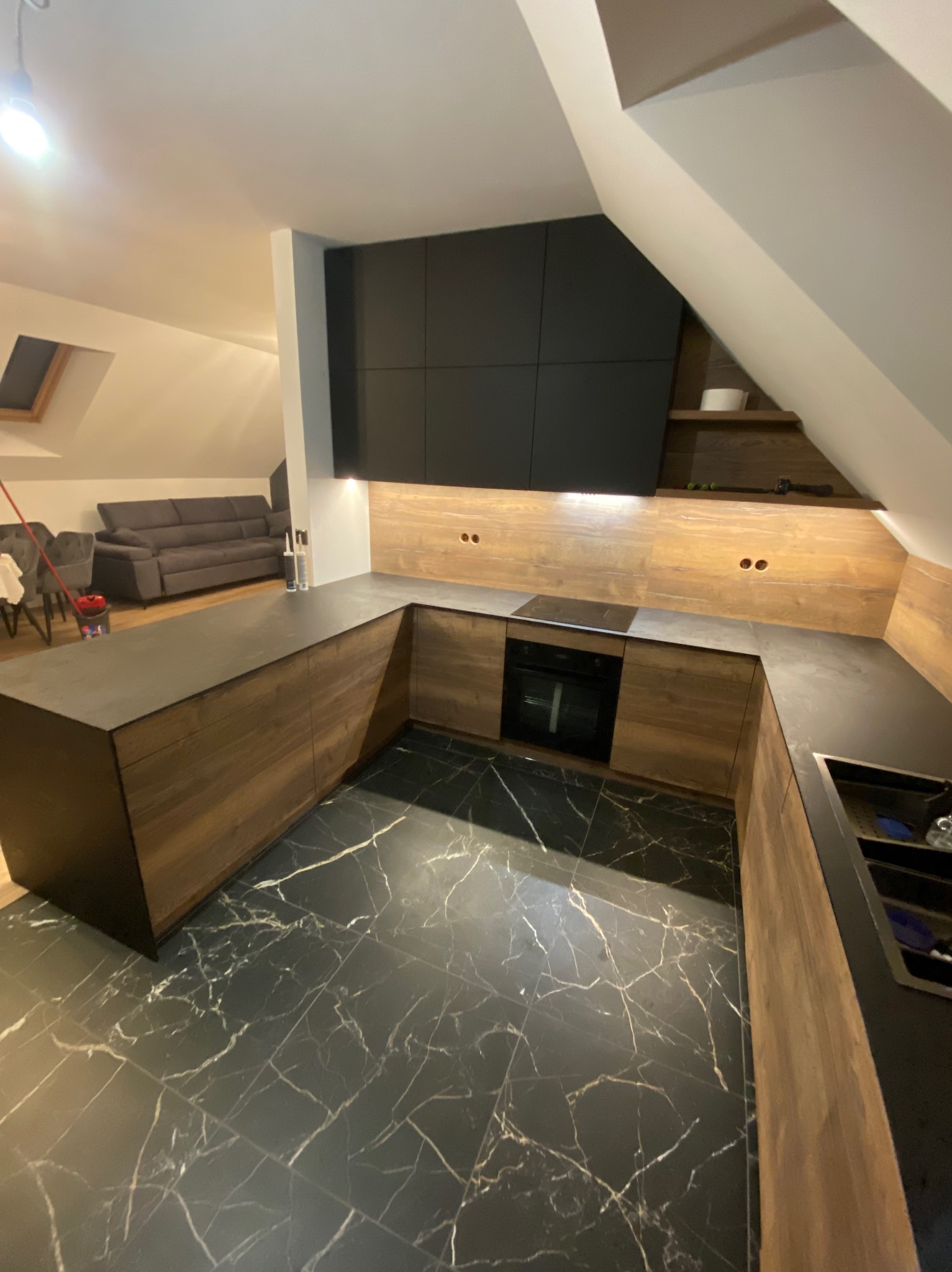 Nowoczesna kuchnia w skosach: czarne szafki górne, drewnopodobne dolne, czarny blat i marmurowa podłoga z widokiem na salon.