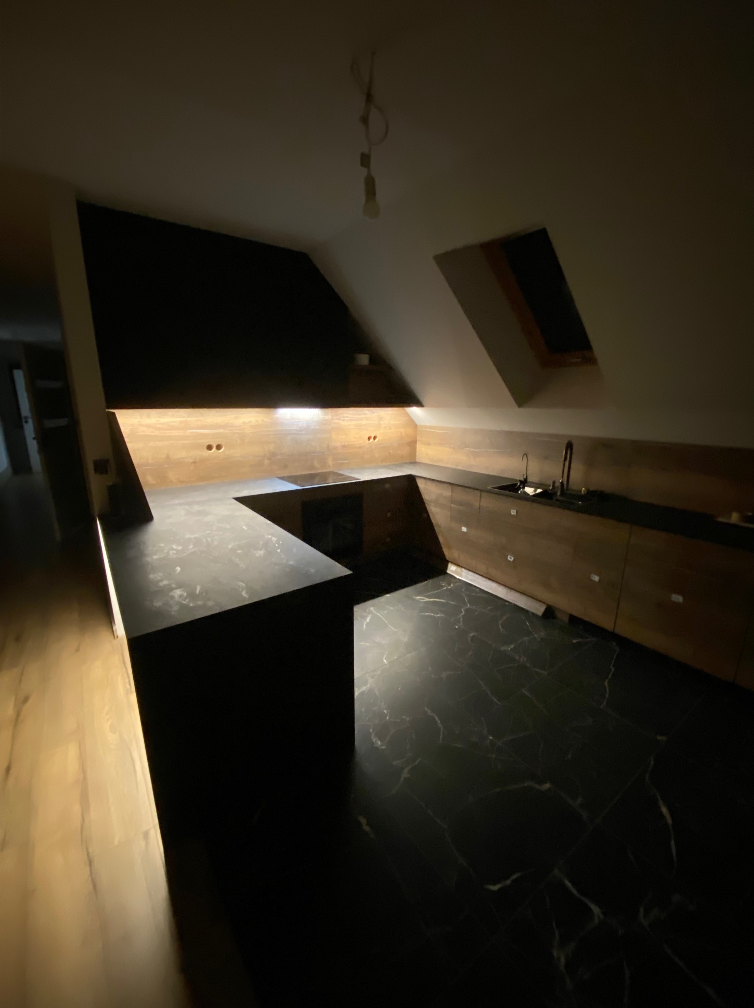 Kuchnia w skosie poddasza z czarnym blatem i marmurową podłogą, oświetlenie podszafkowe.
