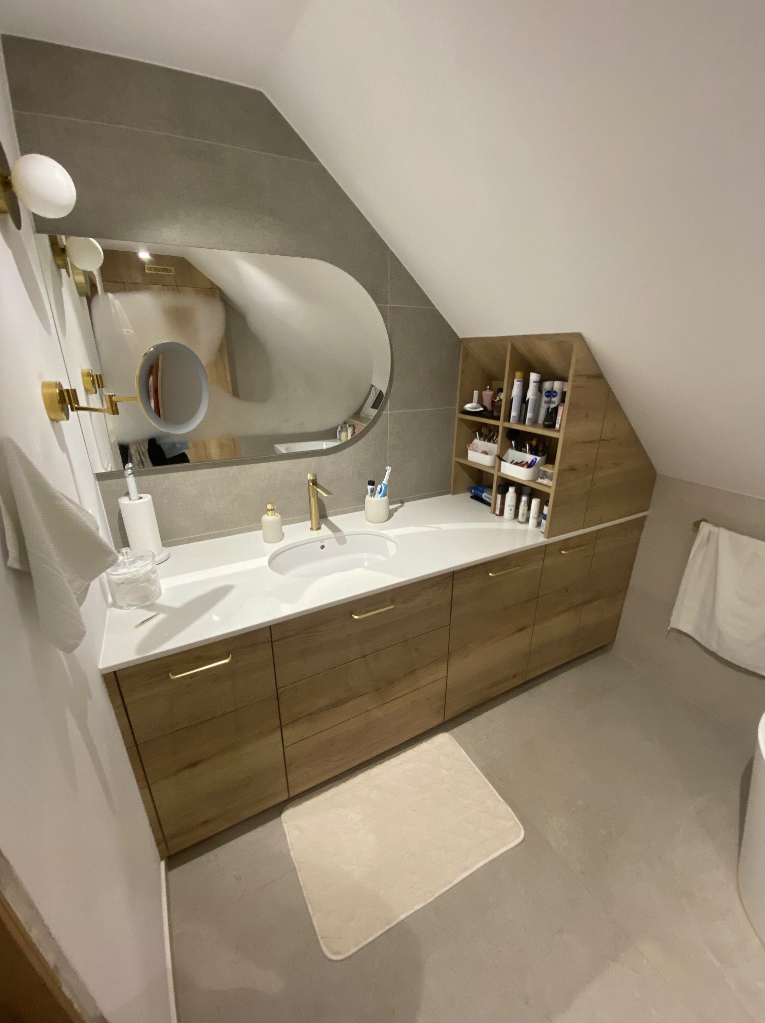 Łazienka na poddaszu z zabudową meblową z drewnianymi frontami, biały blat z umywalką, owalne lustro z podświetleniem, złota armatura, półki na kosmetyki dopasowane do skosu dachu.