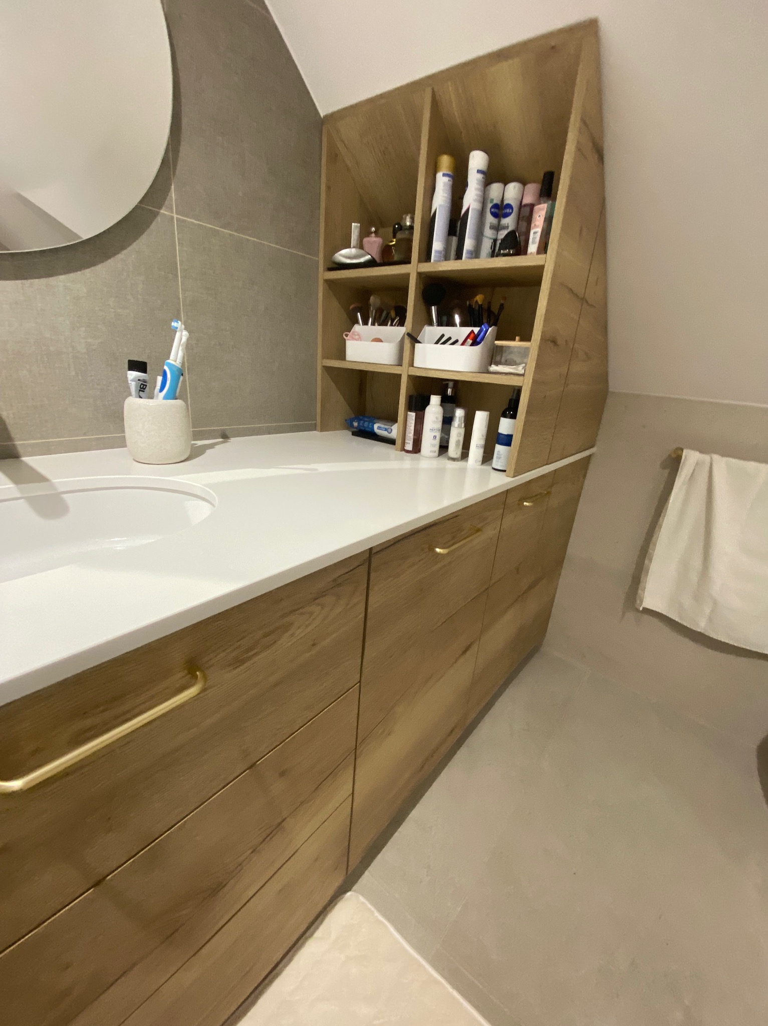 Łazienka z drewnianą szafką pod umywalką z szufladami i otwartymi półkami, na których ustawione są kosmetyki i akcesoria do makijażu, okrągłe lustro nad umywalką, ręcznik na wieszaku z boku.