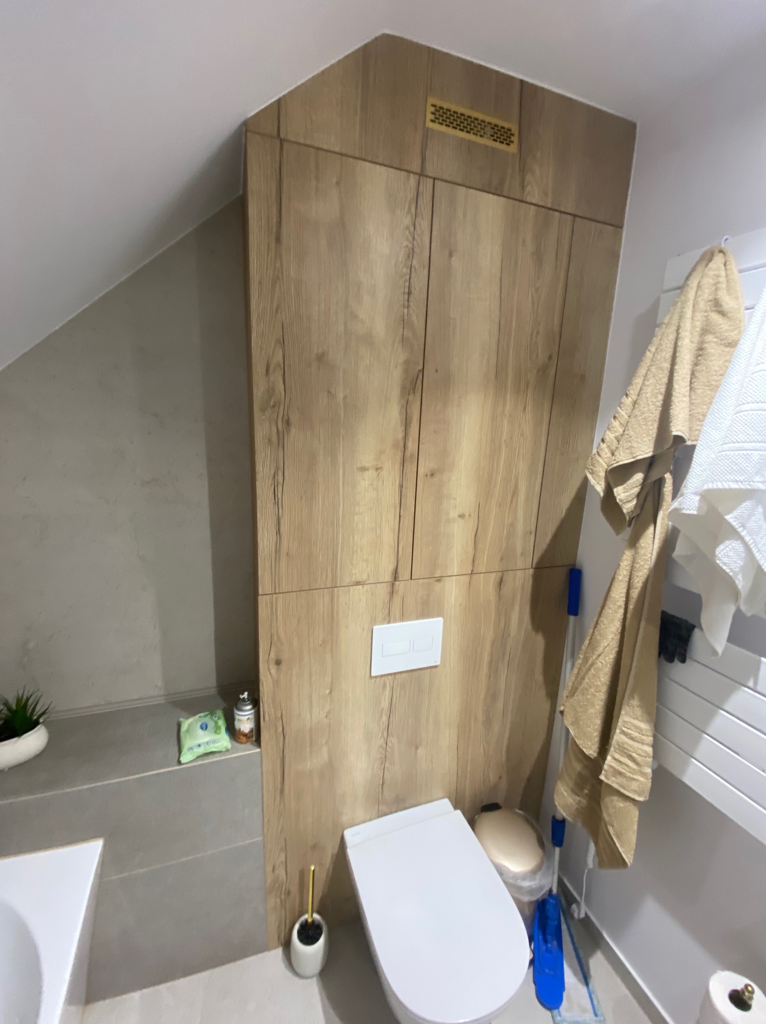 Szafa wnękowa z drewnopodobną okleiną w łazience na poddaszu, zabudowa nad toaletą, widoczny fragment blatu z rośliną i kosmetykami.