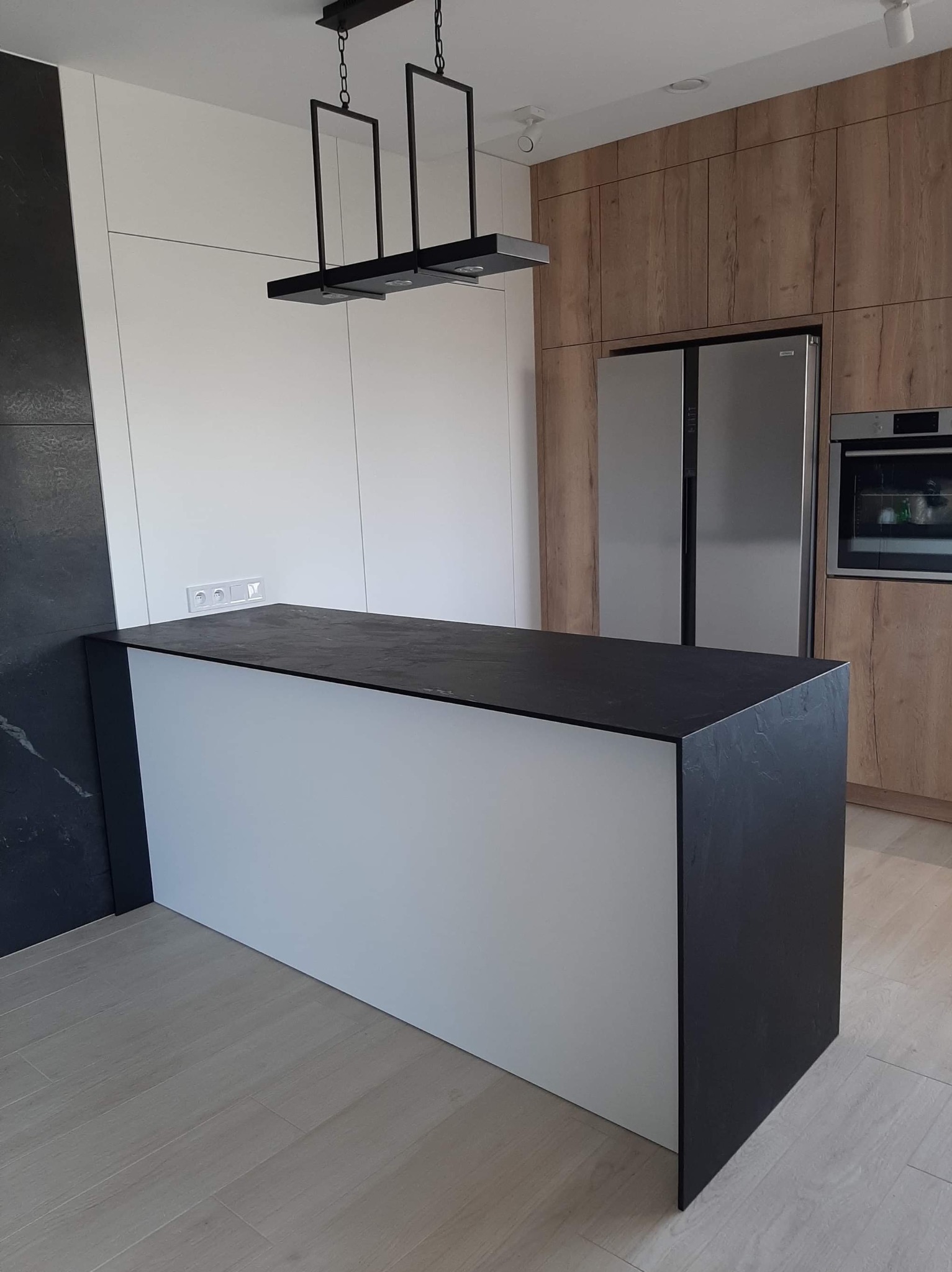 Nowoczesna kuchnia z wyspą o białych frontach i blatem z czarnego kamienia, drewniane szafki w tle, minimalistyczna lampa wisząca.