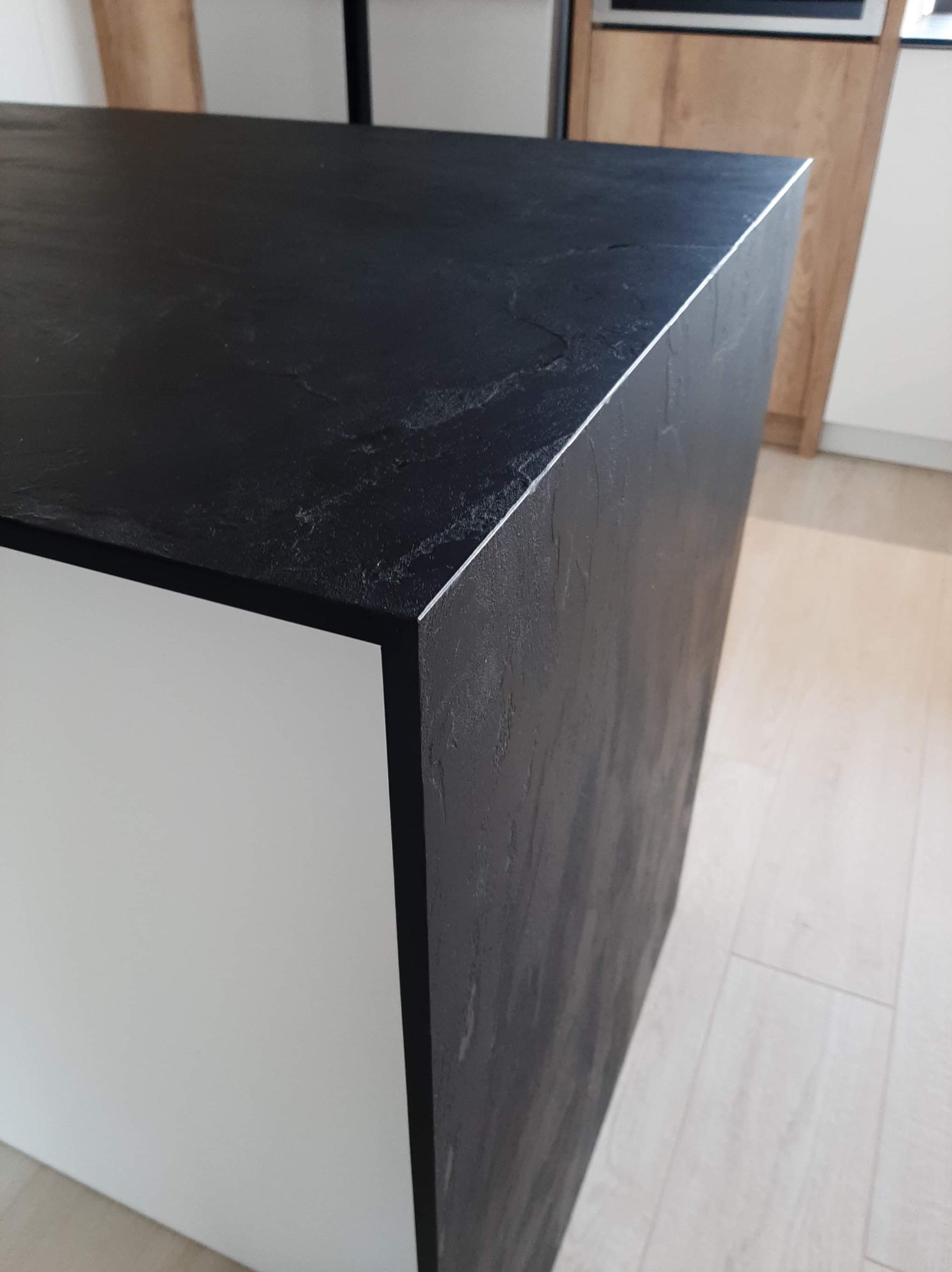 Fragment wyspy kuchennej z blatem z czarnego kamienia o wyraźnej fakturze, połączonego z białym korpusem, widoczny na tle jasnej podłogi i fragmentu zabudowy meblowej.