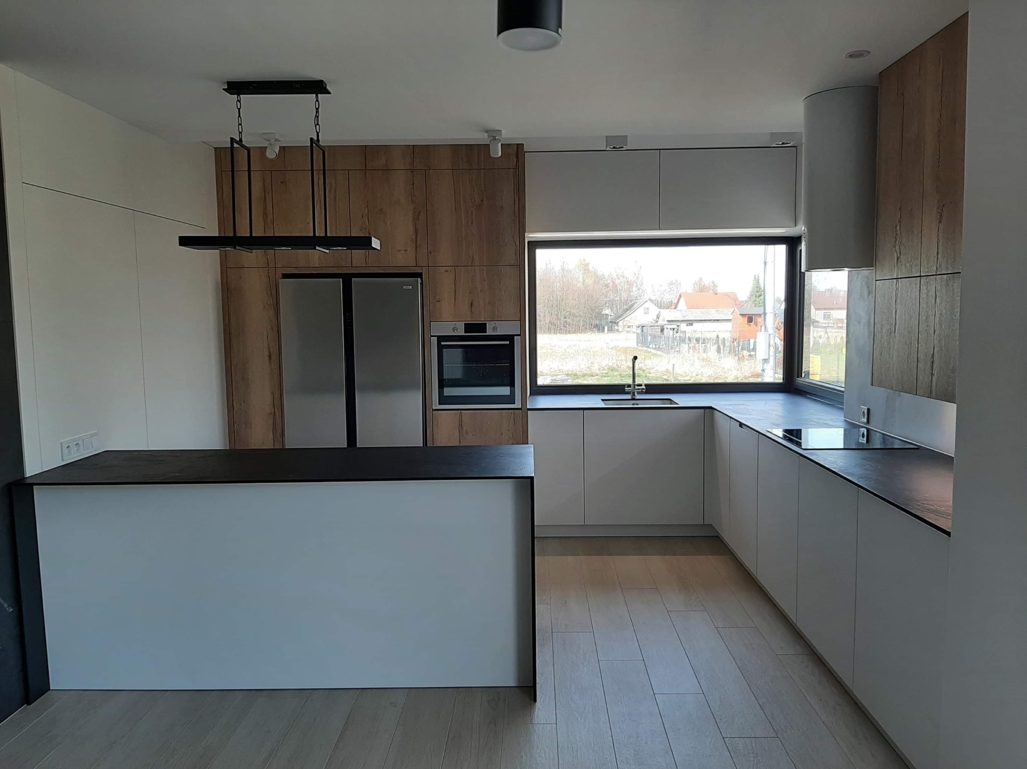 Nowoczesna kuchnia z wyspą, białe i drewnopodobne fronty, czarny blat wyspy, wisząca lampa nad wyspą, zabudowa sięgająca sufitu, widok na domy za oknem.