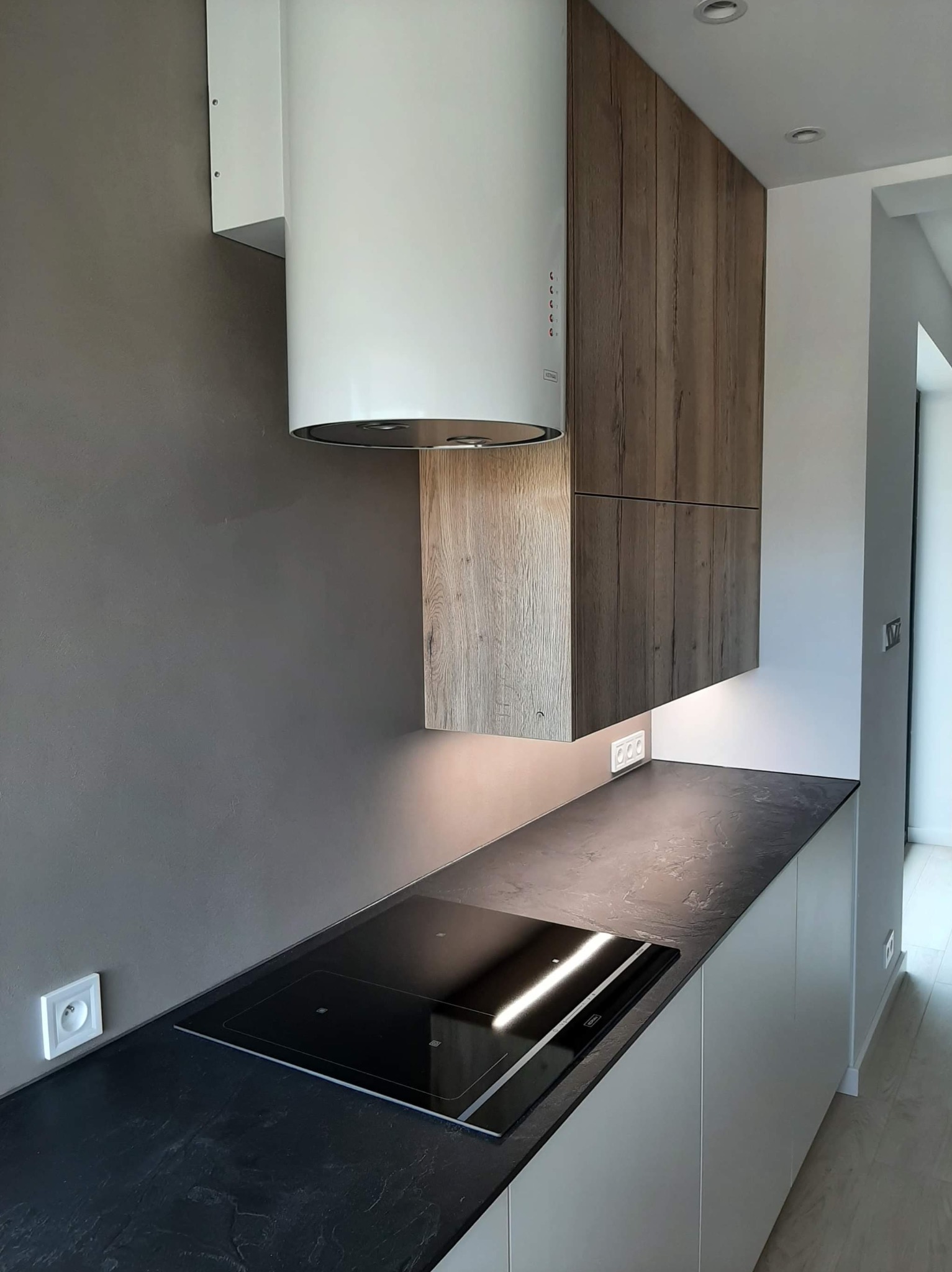 Nowoczesna kuchnia: czarny blat, białe szafki dolne, drewnopodobne szafki wiszące, biały okrągły okap i płyta indukcyjna.