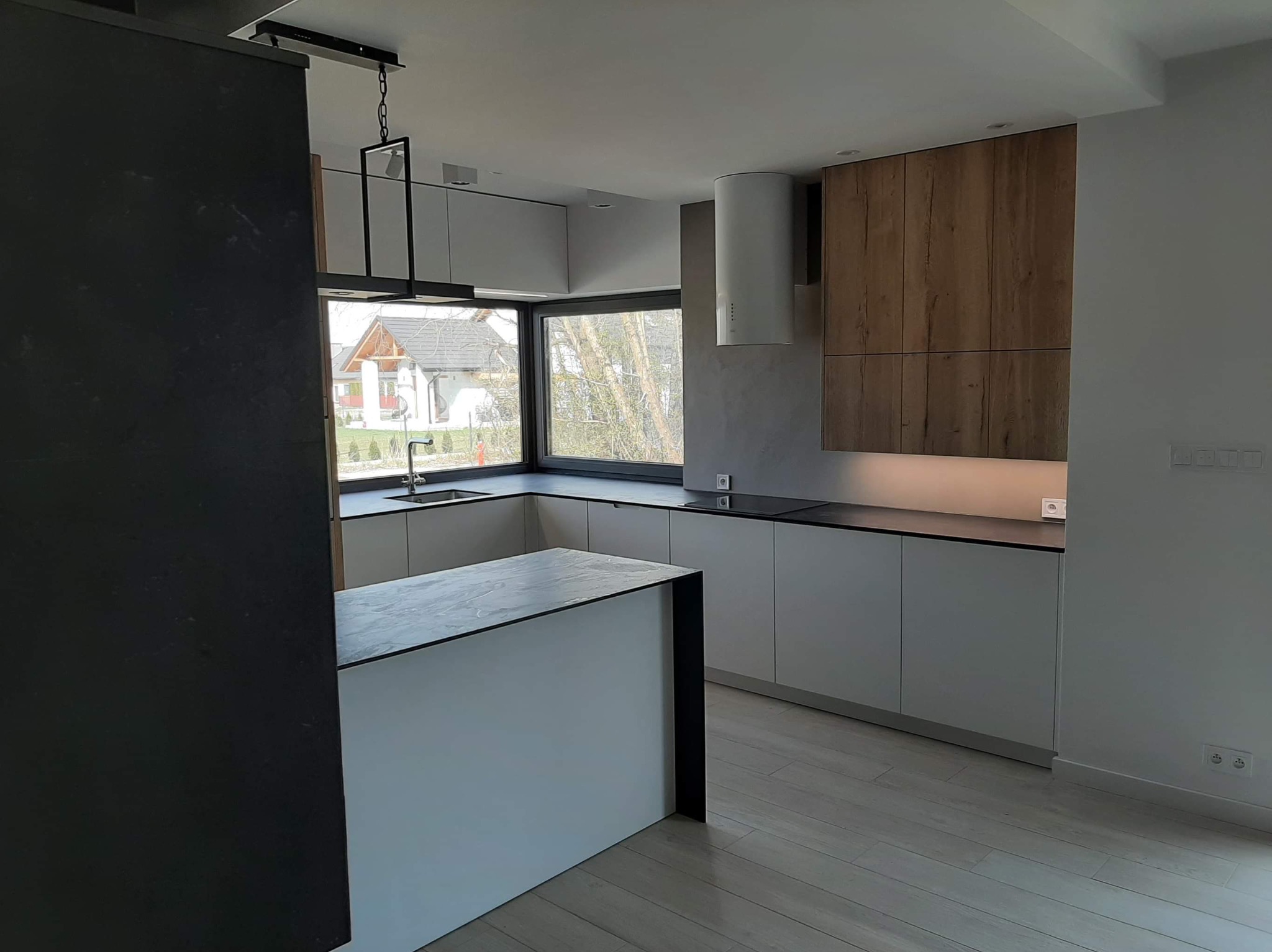 Nowoczesna kuchnia z wyspą w Krakowie: białe fronty szafek dolnych, drewniane szafki górne, czarny blat i okno z widokiem na domy.