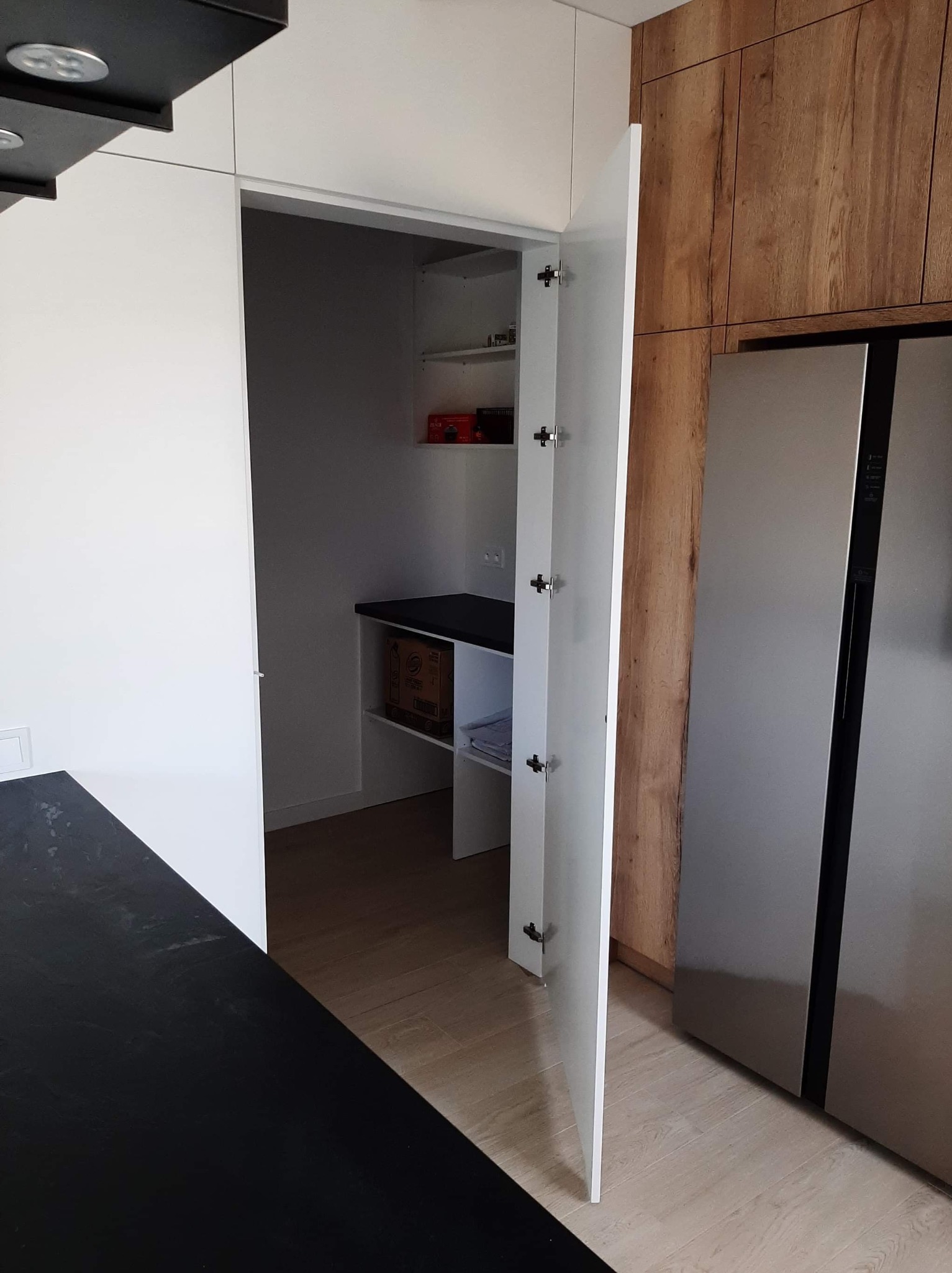 Nowoczesna kuchnia z otwartą szafą wnękową, widoczne półki z artykułami spożywczymi i pudełkami, obok stoi duża lodówka.