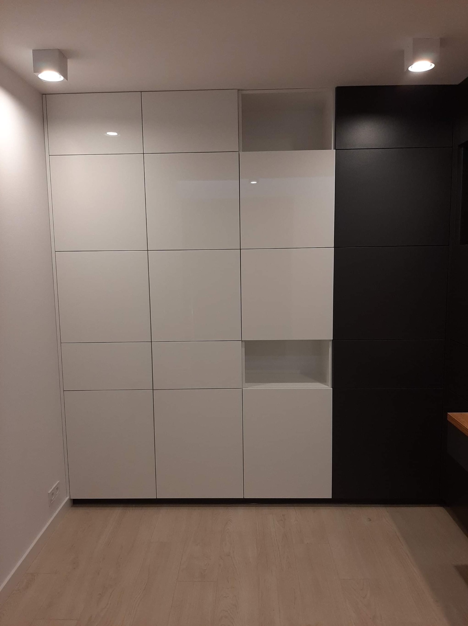 Biało-czarna zabudowa meblowa z błyszczącymi frontami, składająca się z kwadratowych modułów, z otwartymi półkami, oświetlona punktowo, na podłodze panele imitujące drewno.