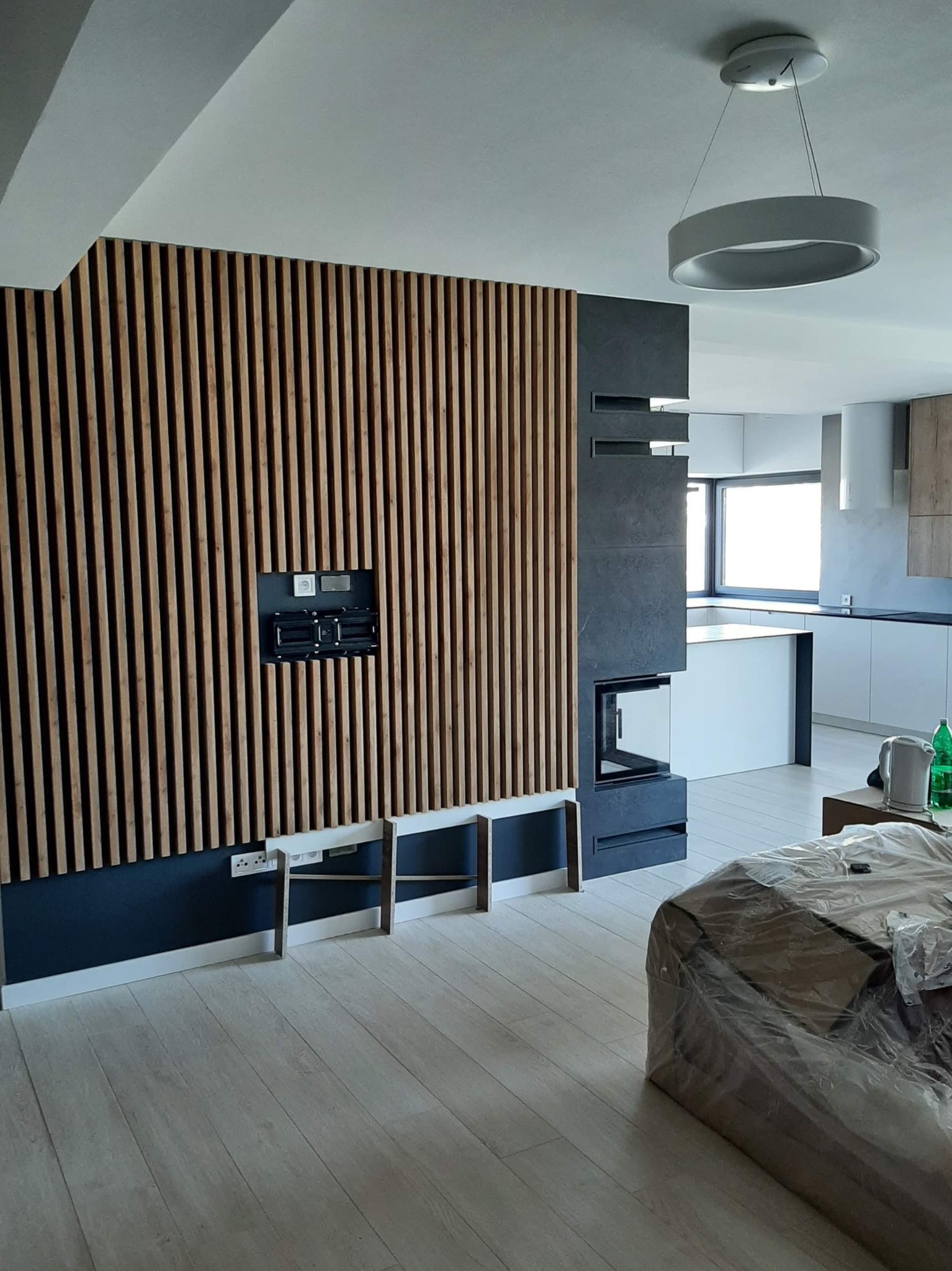 Nowoczesny salon z drewnianą ścianą z pionowych listew, wnęką na telewizor, ciemnoszarym kominkiem i widoczną w tle białą kuchnią z wyspą.