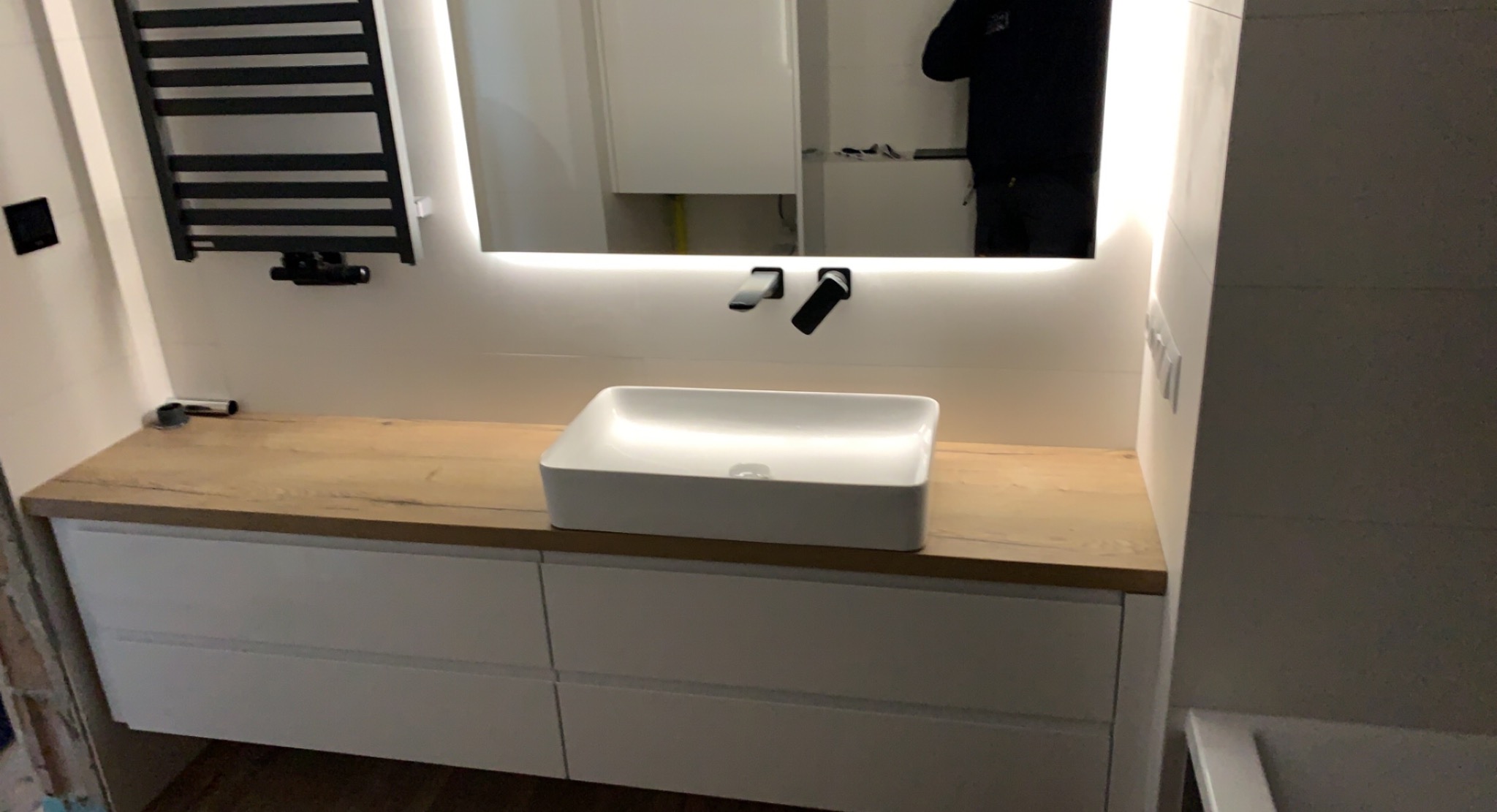 Nowoczesna łazienka z szafką pod umywalką z białymi frontami i blatem imitującym jasne drewno, prostokątna umywalka nablatowa, czarna armatura i podświetlane lustro.