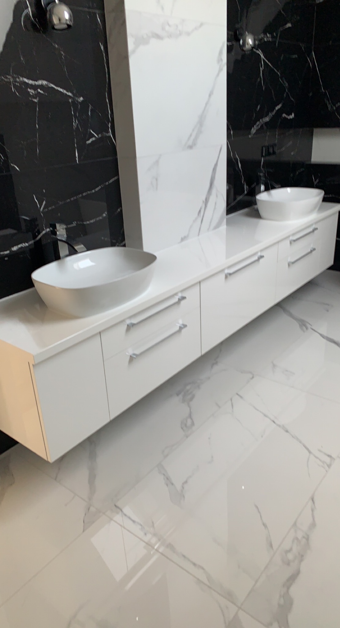 Biała, podwieszana szafka łazienkowa z dwiema owalnymi umywalkami na blacie, minimalistyczne uchwyty, tło z czarno-białych marmurowych płytek, połyskująca podłoga.