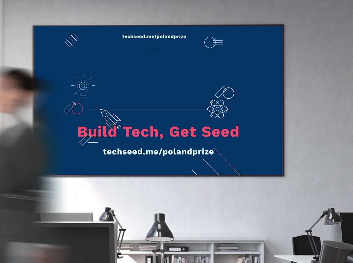 Ekran z grafiką 'Build Tech, Get Seed' i adresem techseed.me/polandprize, wiszący na ścianie w biurze, z widocznym fragmentem osoby i biurka.