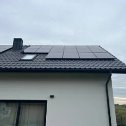 Częściowy widok domu z czarną dachówką i panelami słonecznymi, okno dachowe, komin, biała elewacja, okno z zasłoną.