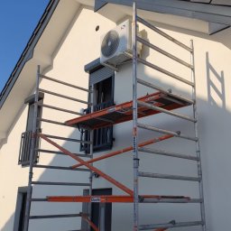 JANCZAR.BIZ DOMINIK JANCZAR - Biały klimatyzator na ścianie domu, zamontowany nad oknem z roletą, widoczny ze rusztowania na tle błękitnego nieba.