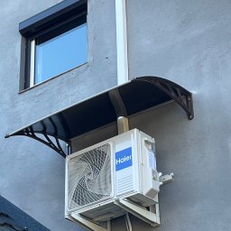 JANCZAR.BIZ DOMINIK JANCZAR - Zamontowana jednostka zewnętrzna klimatyzacji Haier na szarej elewacji budynku, osłonięta daszkiem. Widoczny fragment okna z roletą i rura spustowa.