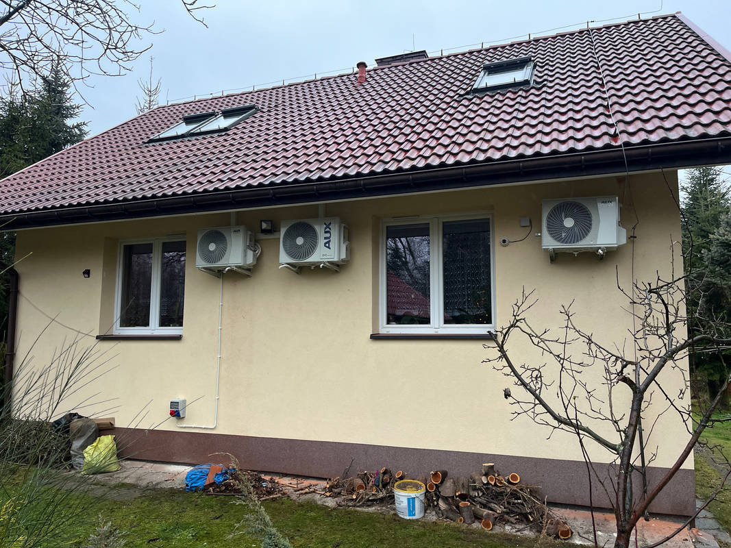 Dom jednorodzinny z trzema zamontowanymi klimatyzatorami na elewacji, widoczne okna dachowe i przycięte gałęzie drzew na trawniku.