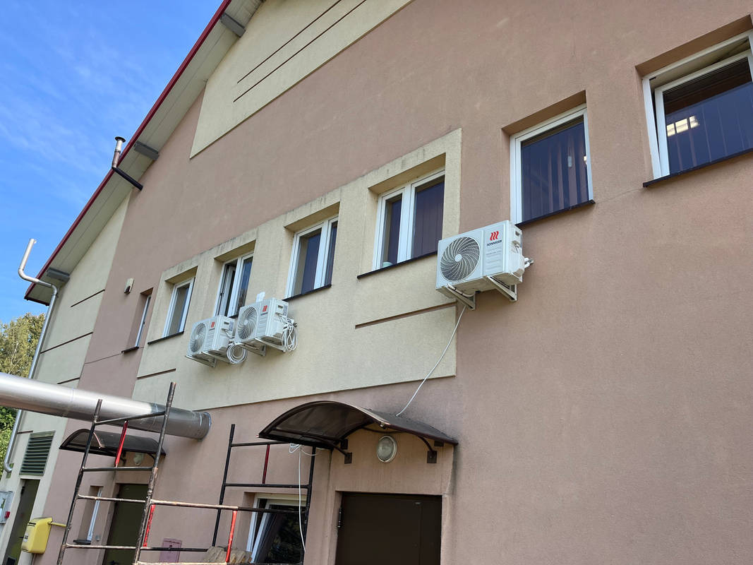 Zewnętrzne jednostki klimatyzacji zamontowane na elewacji budynku biurowego, widoczne okna i zadaszenia wejść.