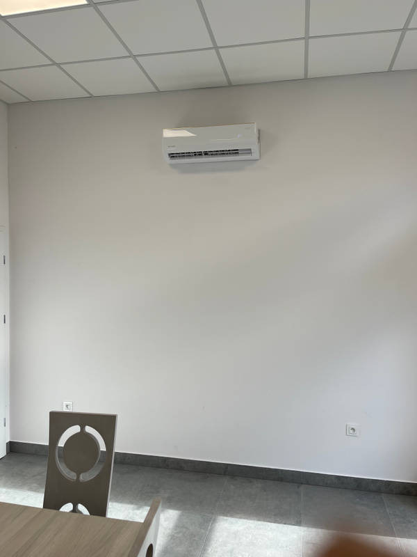 Biała jednostka klimatyzacji zamontowana na ścianie w jasnym pomieszczeniu biurowym, widoczny fragment krzesła i gniazdko elektryczne.