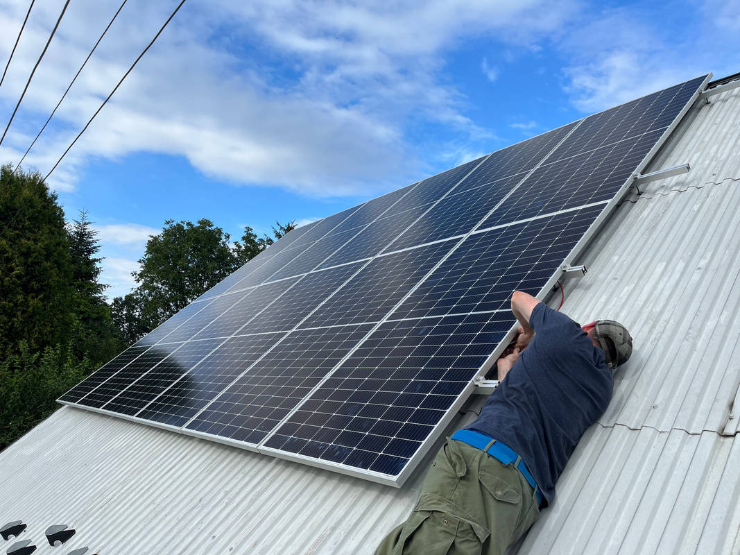 Montaż paneli fotowoltaicznych na blaszanym dachu budynku jednorodzinnego, widoczne narzędzia i instalator w trakcie pracy, błękitne niebo w tle.