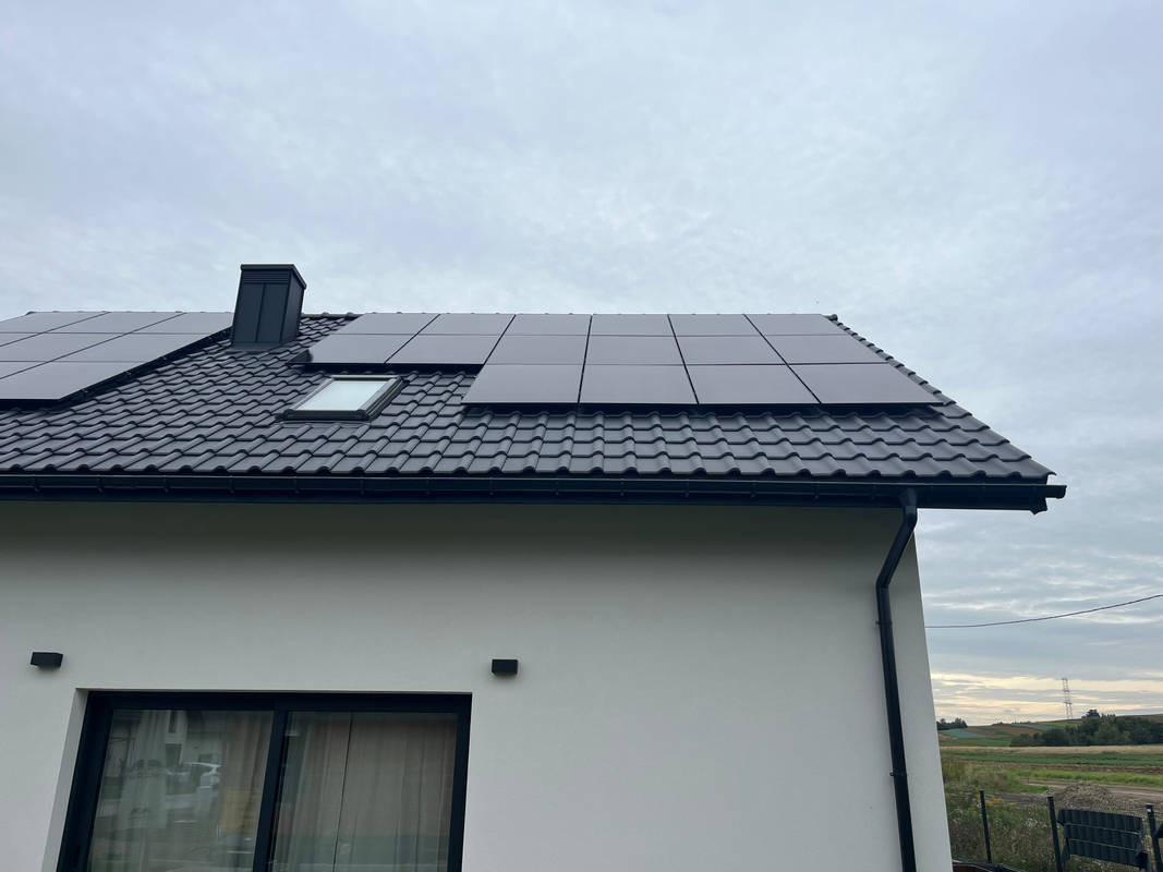 Częściowy widok domu z czarną dachówką i panelami słonecznymi, okno dachowe, komin, biała elewacja, okno z zasłoną.