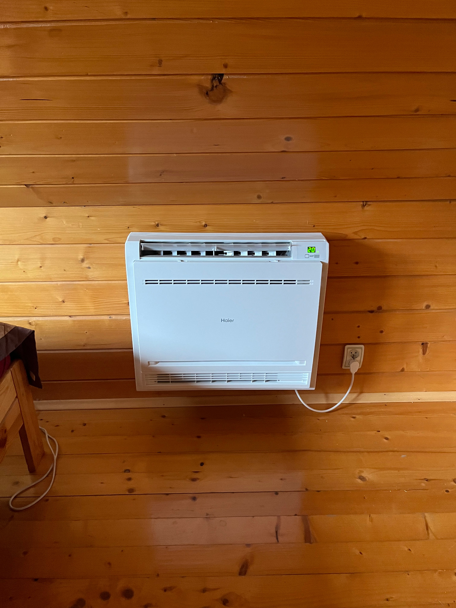 Biały klimatyzator Haier zamontowany na ścianie z drewnianych paneli w ciepłym odcieniu, widoczny kabel zasilający i gniazdko. Wyświetlacz pokazuje temperaturę 20 stopni.