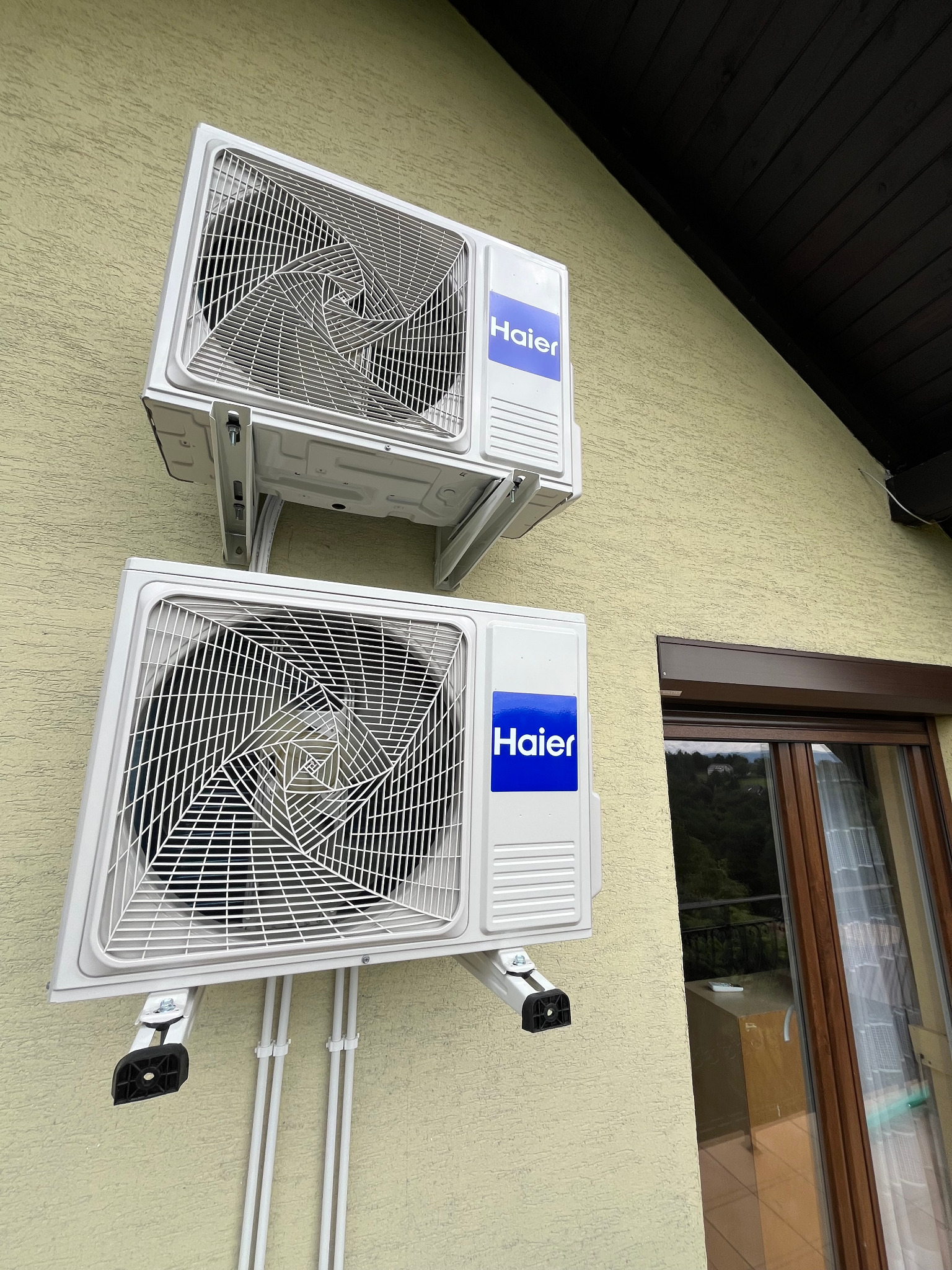 Dwa białe klimatyzatory Haier zamontowane na elewacji budynku z oknem. Widoczne przewody instalacyjne i elementy mocujące. Ujęcie z dołu, lekko z prawej strony.