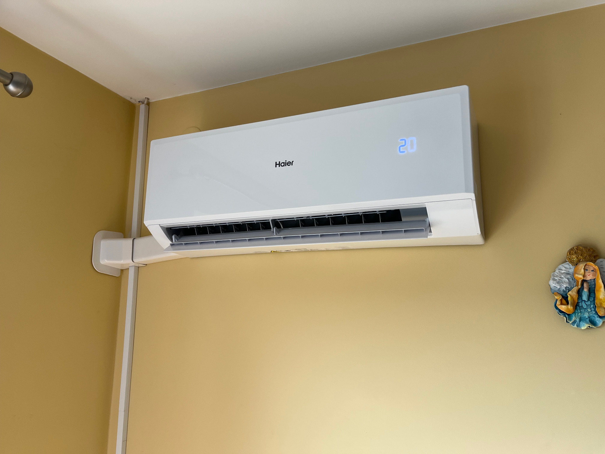 Biały klimatyzator Haier z wyświetlaczem temperatury (20°C) zamontowany na żółtej ścianie w pomieszczeniu. Widoczne rury instalacyjne i dekoracja ścienna.