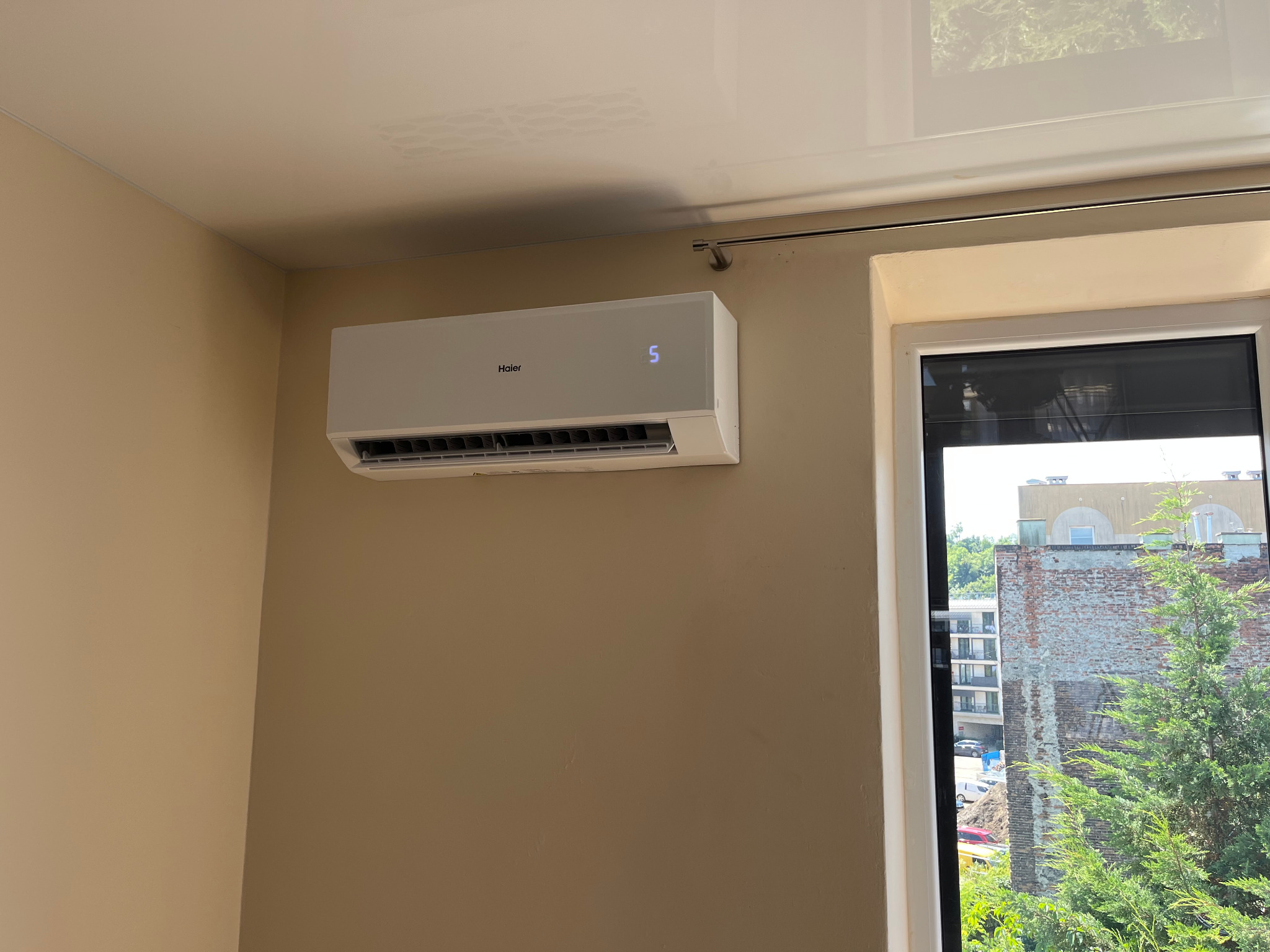 Biały klimatyzator Haier z wyświetlaczem temperatury zamontowany na ścianie w pomieszczeniu w Stronie. Widok z okna na zieleń i budynki.