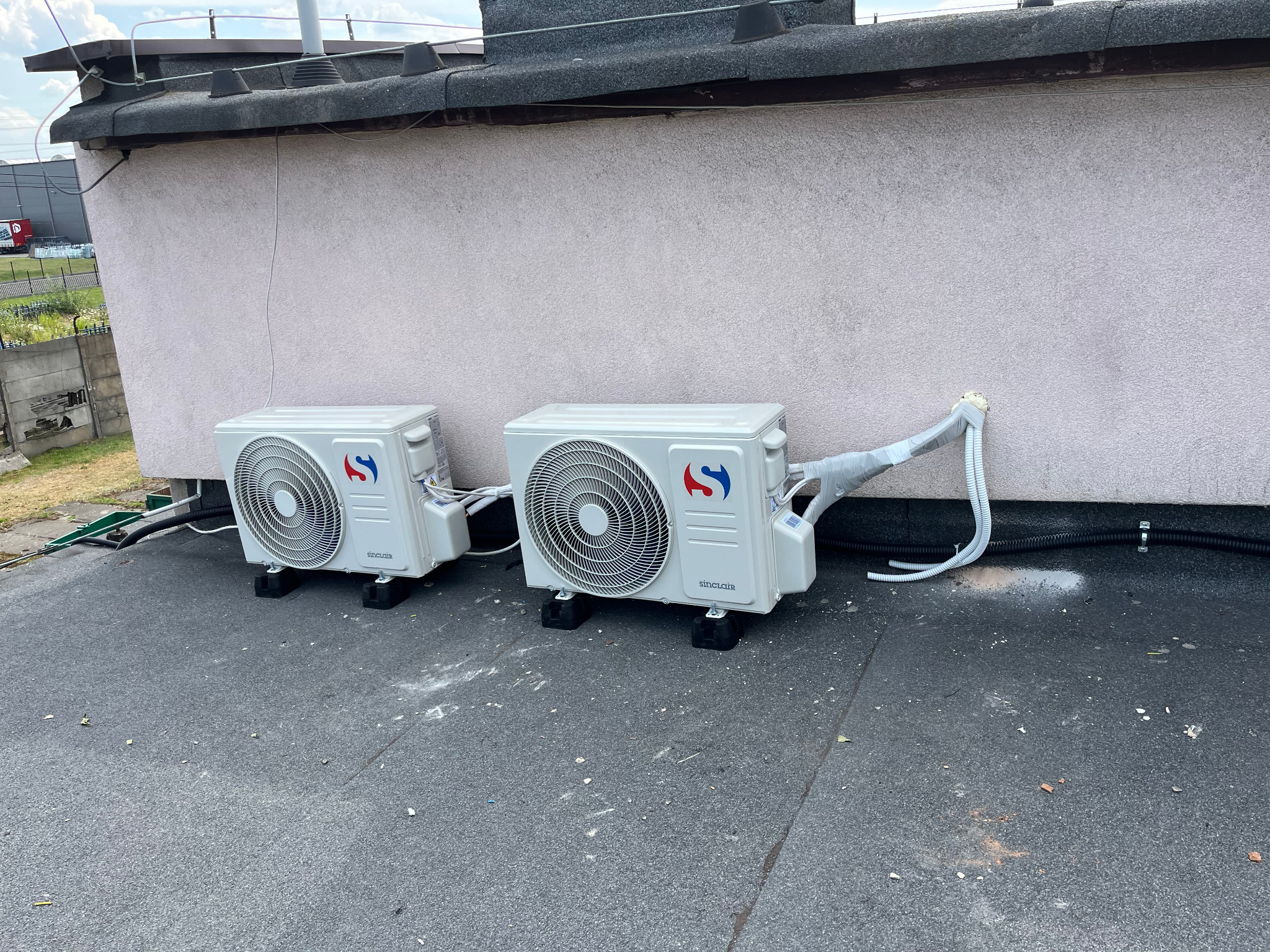 Dwa klimatyzatory Sinclair zamontowane na dachu budynku, widoczne przewody i elementy instalacji. Ściana budynku w tle, dach pokryty papą.