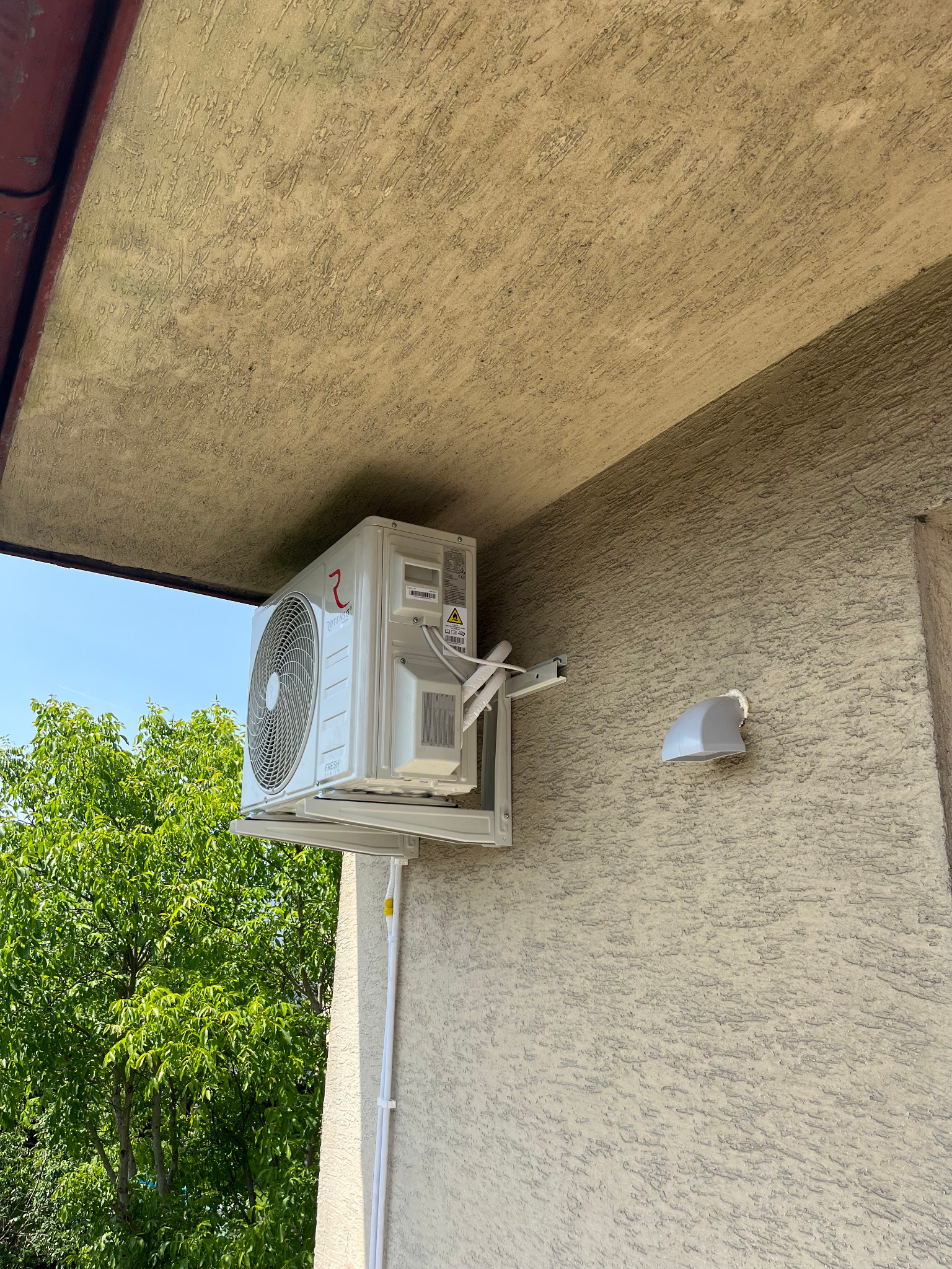 Biała jednostka zewnętrzna klimatyzacji Rotenso zamontowana na elewacji budynku z widocznymi przewodami i elementami montażowymi. W tle zieleń drzew.
