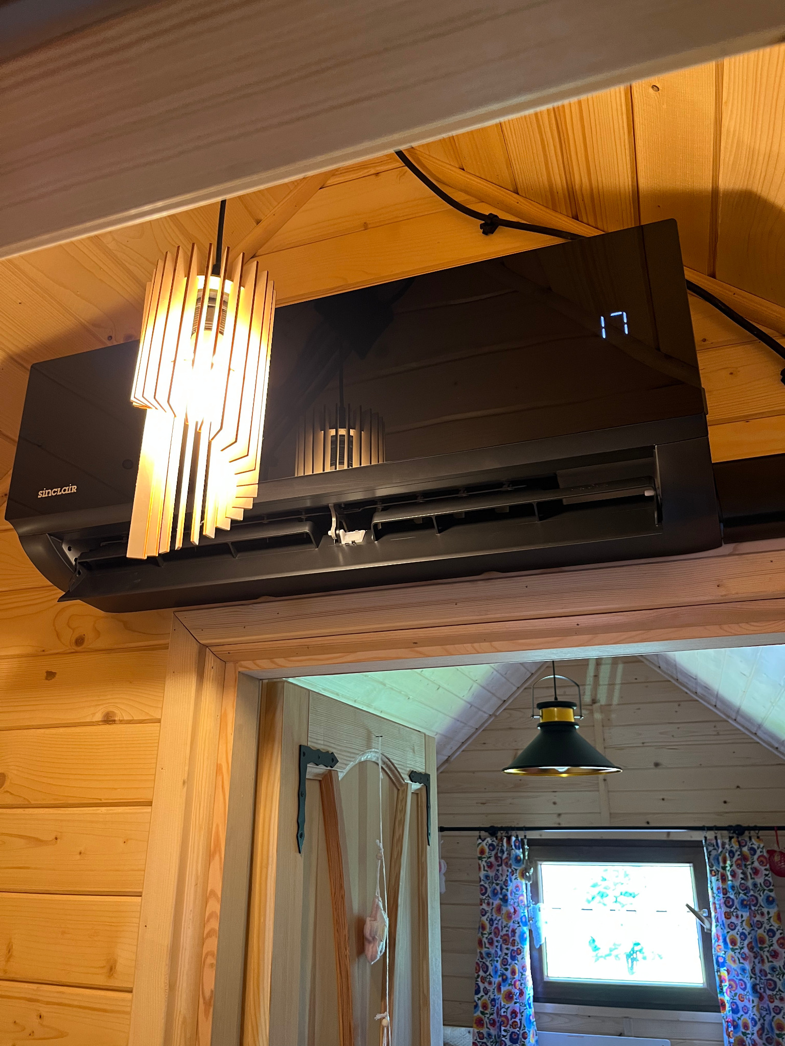 Czarny klimatyzator Sinclair zamontowany w drewnianym domku, oświetlony lampą z drewnianych listewek. Widoczny fragment okna z kolorowymi zasłonami.