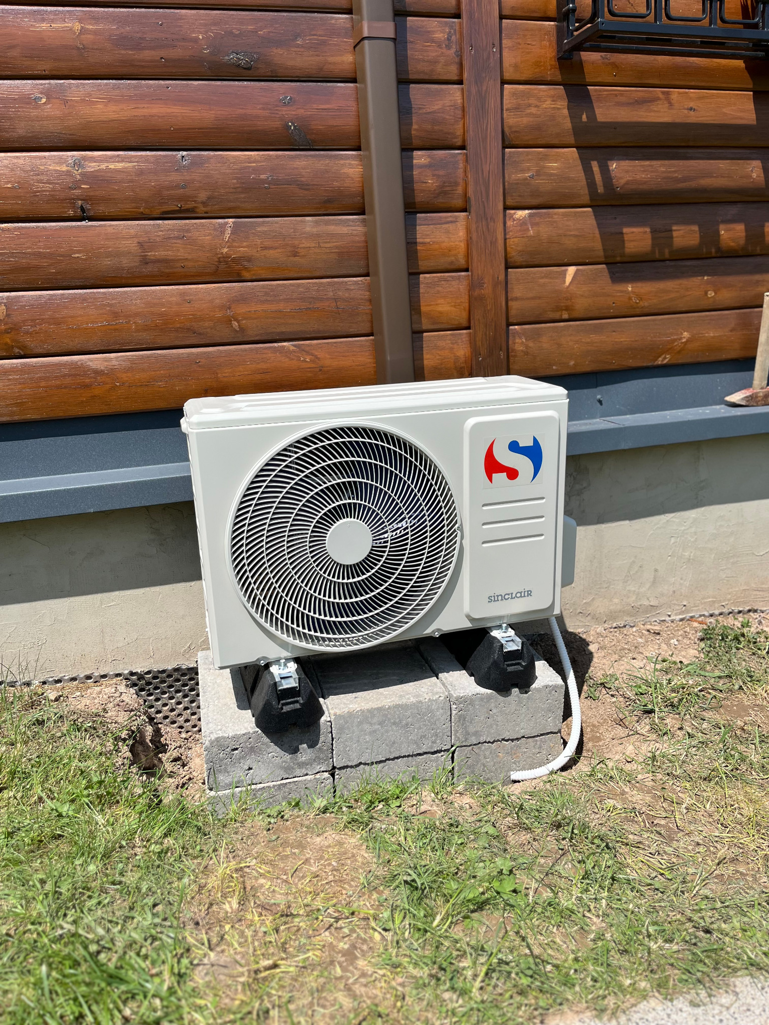 Biała jednostka zewnętrzna klimatyzacji Sinclair, zamontowana na betonowych bloczkach przy drewnianej ścianie budynku. Widoczny kabel zasilający i trawa.