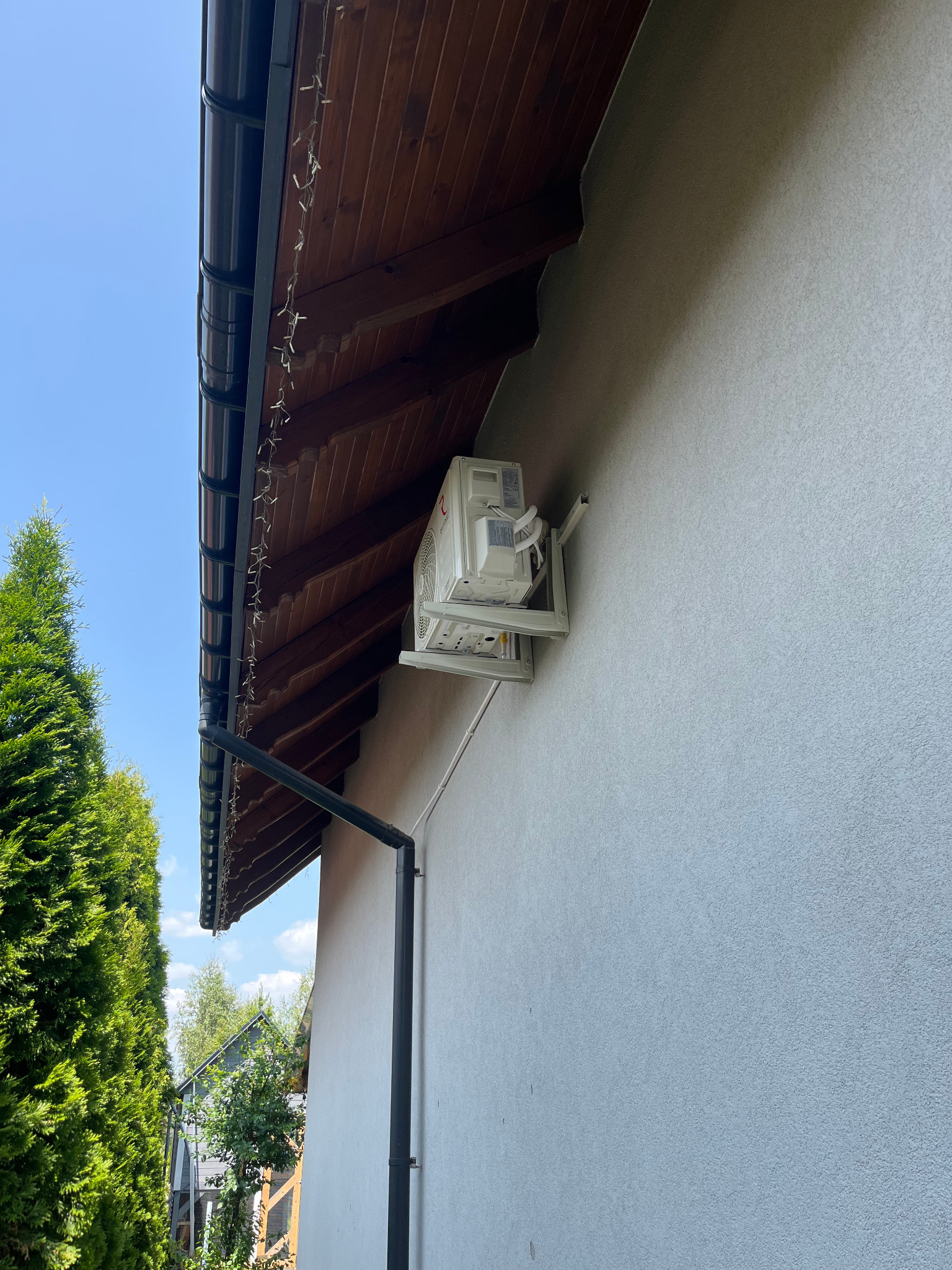 Biała jednostka zewnętrzna klimatyzacji zamontowana na ścianie budynku z szarą elewacją, widok z dołu, z fragmentem dachu i zieleni w tle.