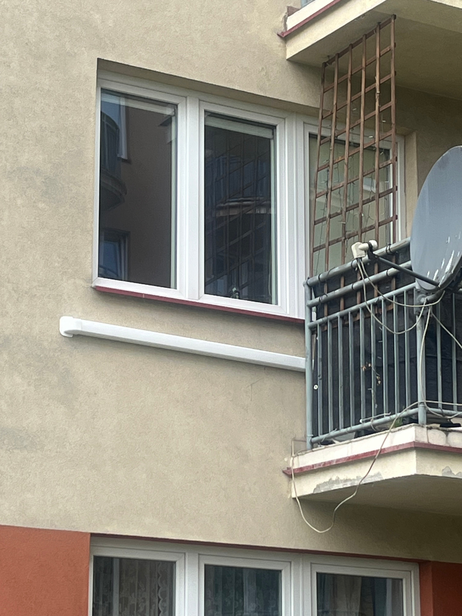 Biała rura klimatyzacji na elewacji budynku, widoczna z zewnątrz, obok okna i balkonu z kratą oraz anteną satelitarną. Szara elewacja, częściowo pomalowana na pomarańczowo.