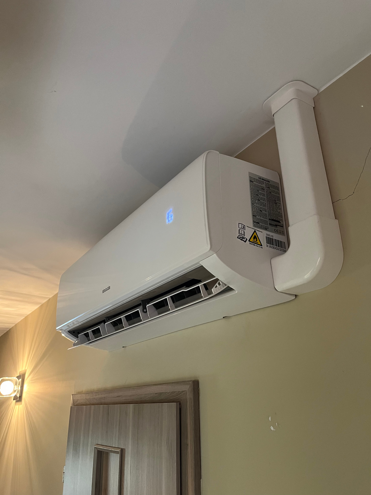 Biały klimatyzator Sinclair zamontowany na ścianie nad drzwiami, z widoczną instalacją odprowadzającą. Na ścianie widoczna lampa i ślady po naprawach.