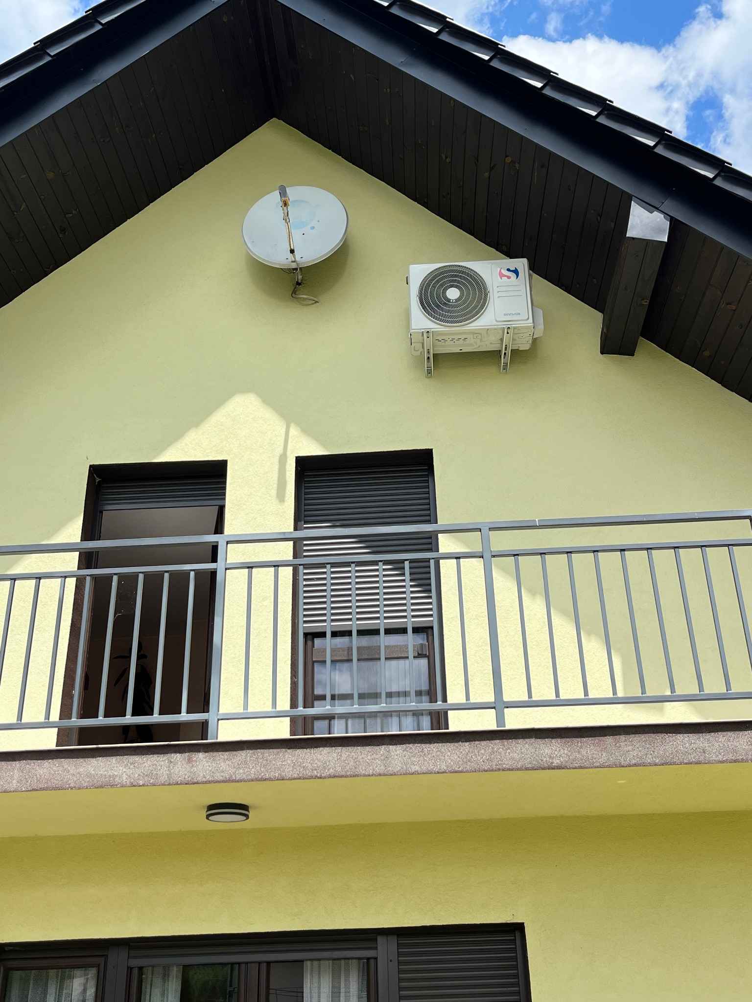 Żółta elewacja domu z balkonem, na której zamontowano klimatyzator marki Sencair i antenę satelitarną. Widoczne okna z roletami i ciemne drewniane elementy dachu.