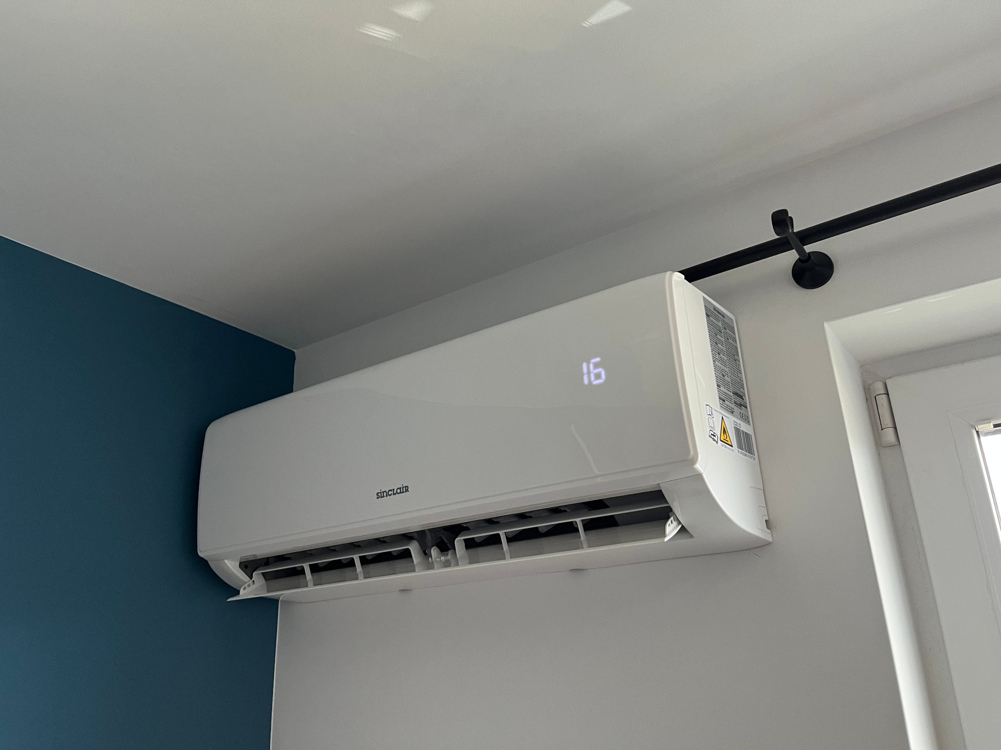 Biały klimatyzator Sinclair na ścianie, ustawiony na 16 stopni, widoczny fragment niebieskiej ściany i karnisz. Nowoczesny design, wnętrze domu.