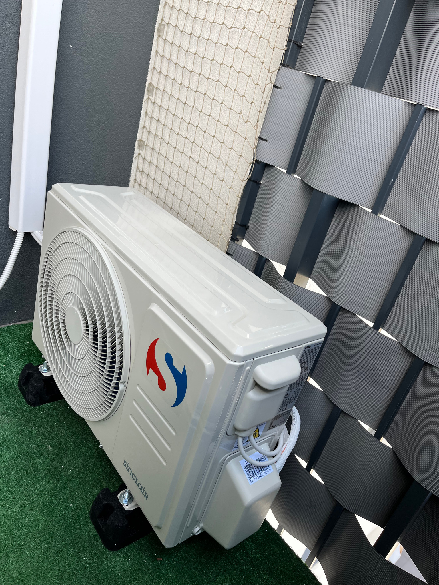 Biała jednostka zewnętrzna klimatyzacji Sinclair na zielonej powierzchni, z widocznymi elementami montażowymi i szarą ażurową osłoną w tle.