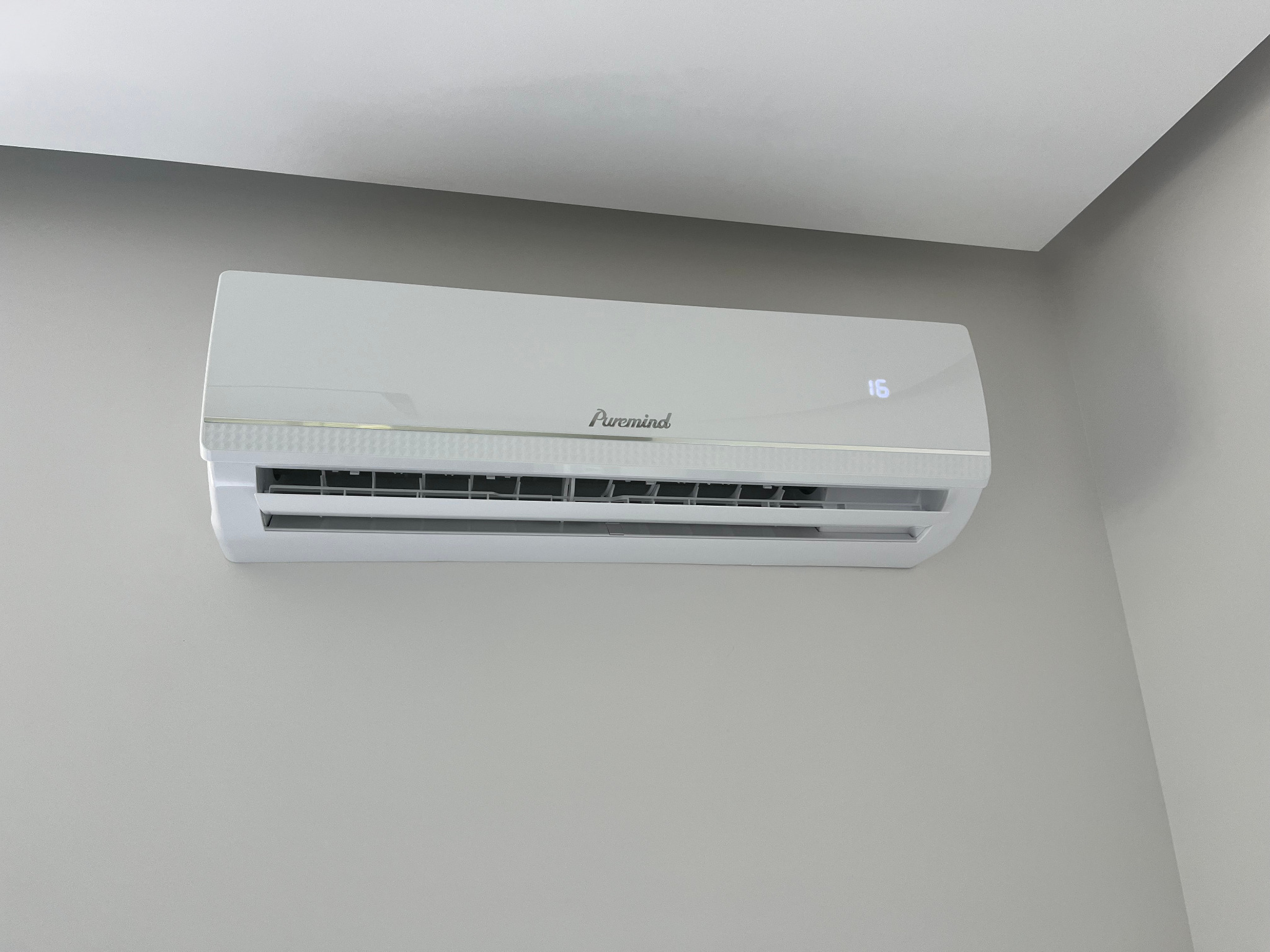 Biała klimatyzacja Puremind zamontowana na szarej ścianie, widoczny wyświetlacz z temperaturą 16 stopni, nowoczesny design, wnętrze pomieszczenia.