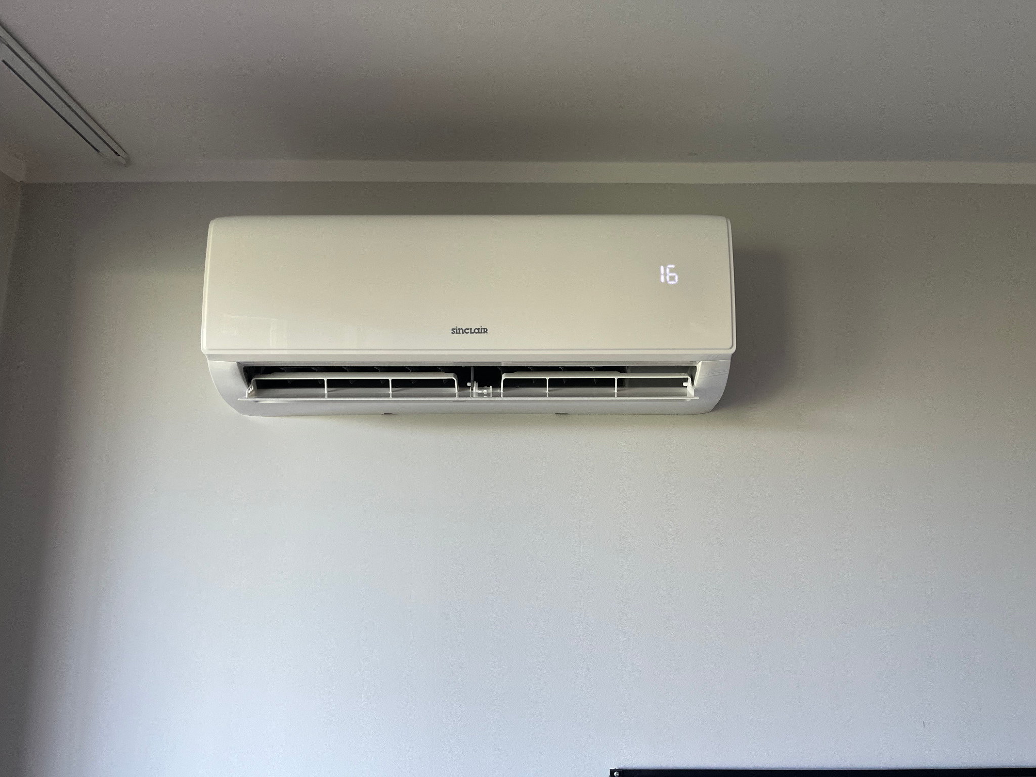 Biała klimatyzacja Sinclair zamontowana na jasnej ścianie. Na wyświetlaczu widoczna temperatura 16 stopni. Widok z dołu, lekko z prawej strony.