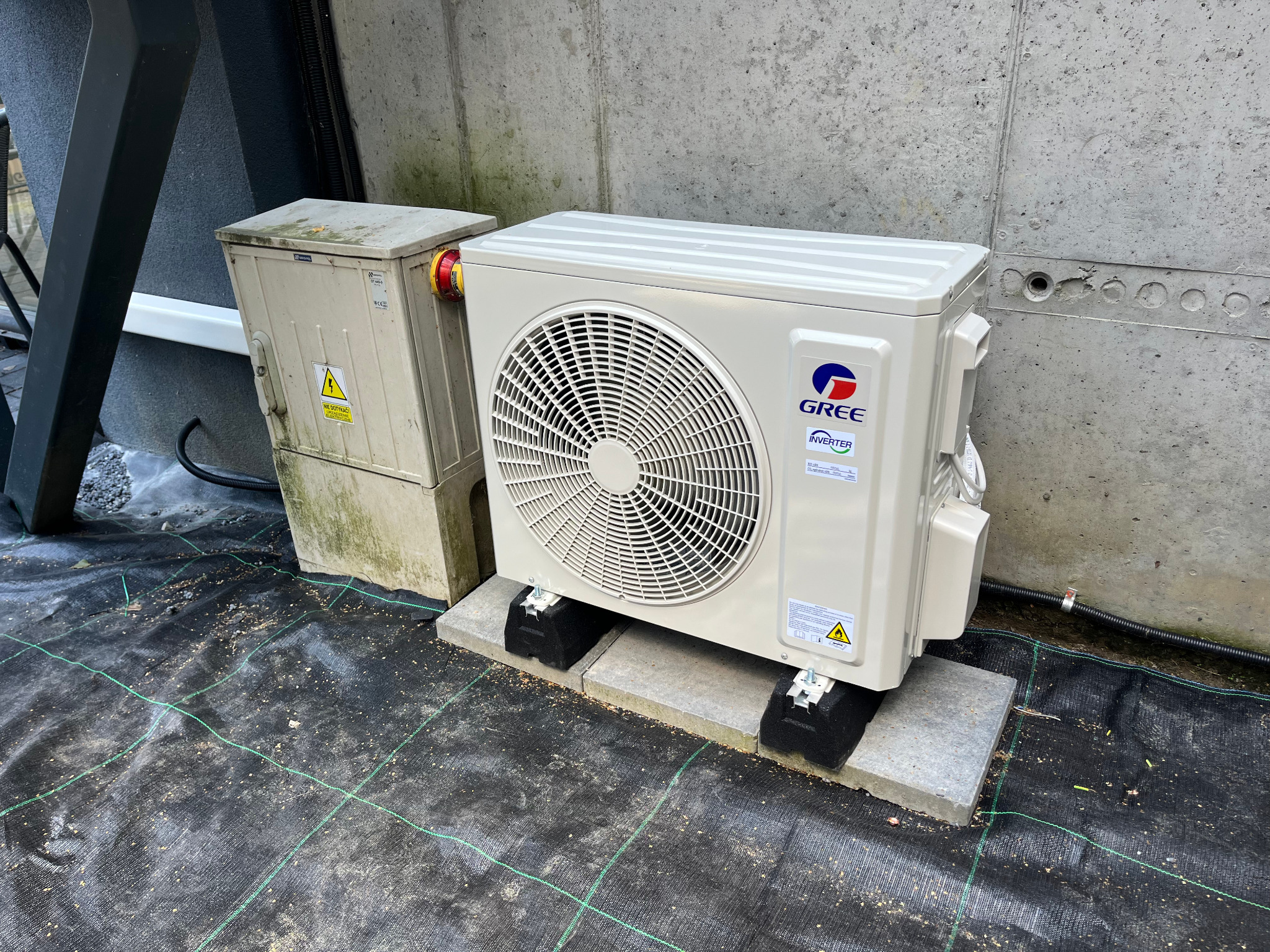 Zewnętrzna jednostka klimatyzacji Gree, zamontowana na betonowych bloczkach, z widoczną skrzynką elektryczną obok, na tle betonowej ściany i czarnej maty.