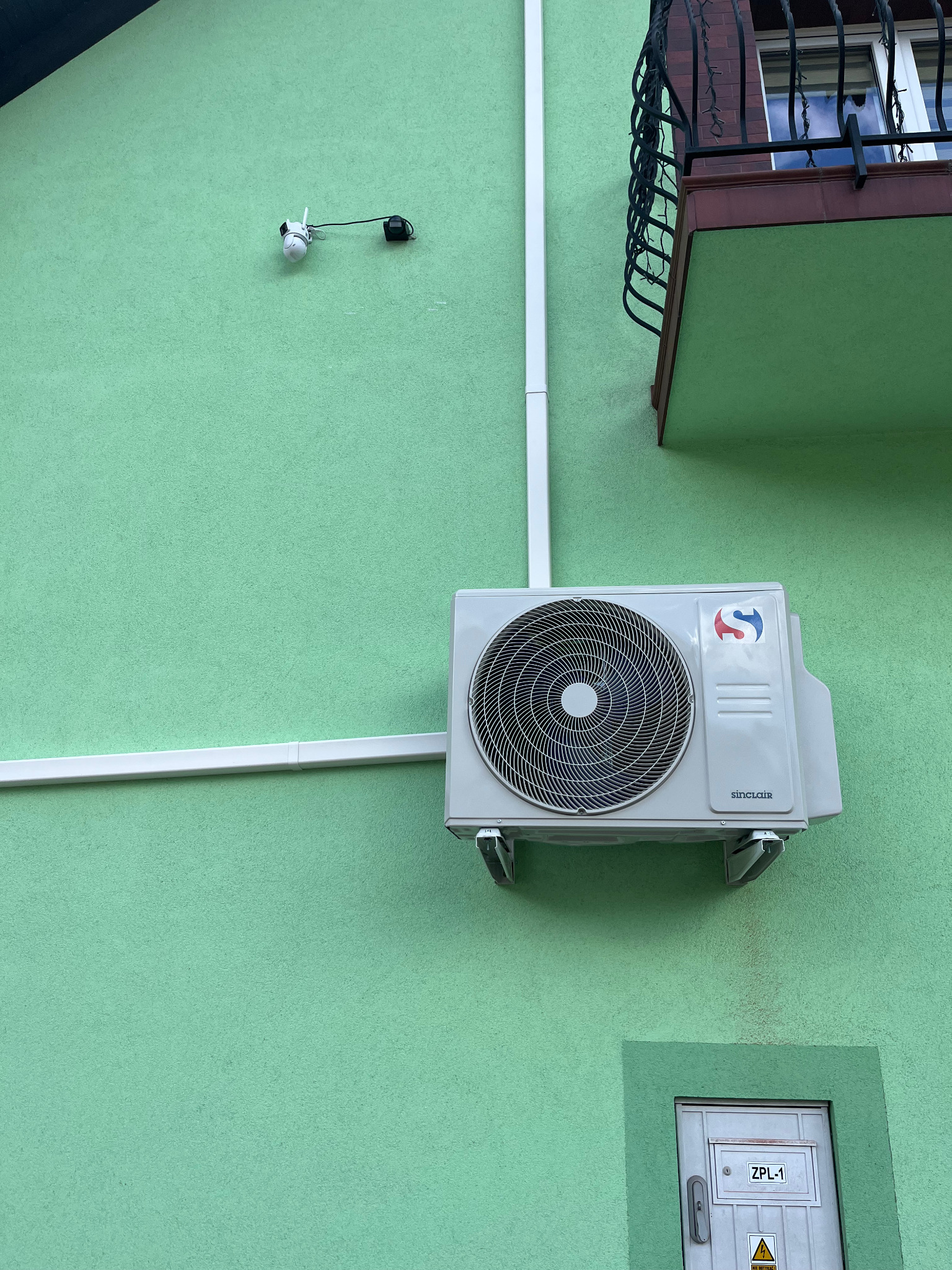 Zamontowana klimatyzacja Sinclair na zielonej ścianie budynku w Stronie. Widoczne rury instalacyjne i balkon z ozdobną balustradą. Ujęcie z dołu.