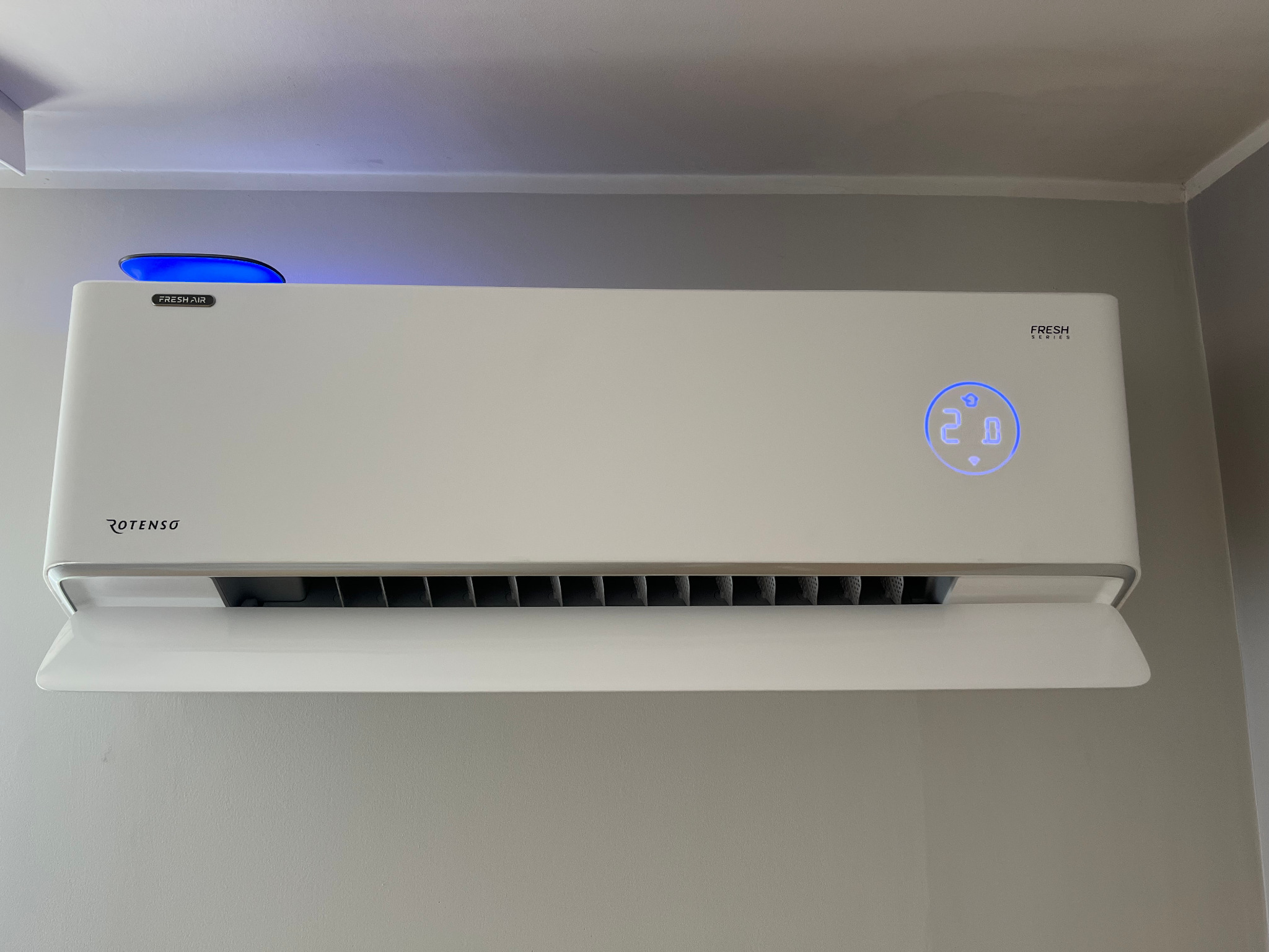 Biała klimatyzacja Rotenso z niebieskim podświetleniem, zamontowana na szarej ścianie. Widoczny wyświetlacz temperatury i logo Fresh Air.