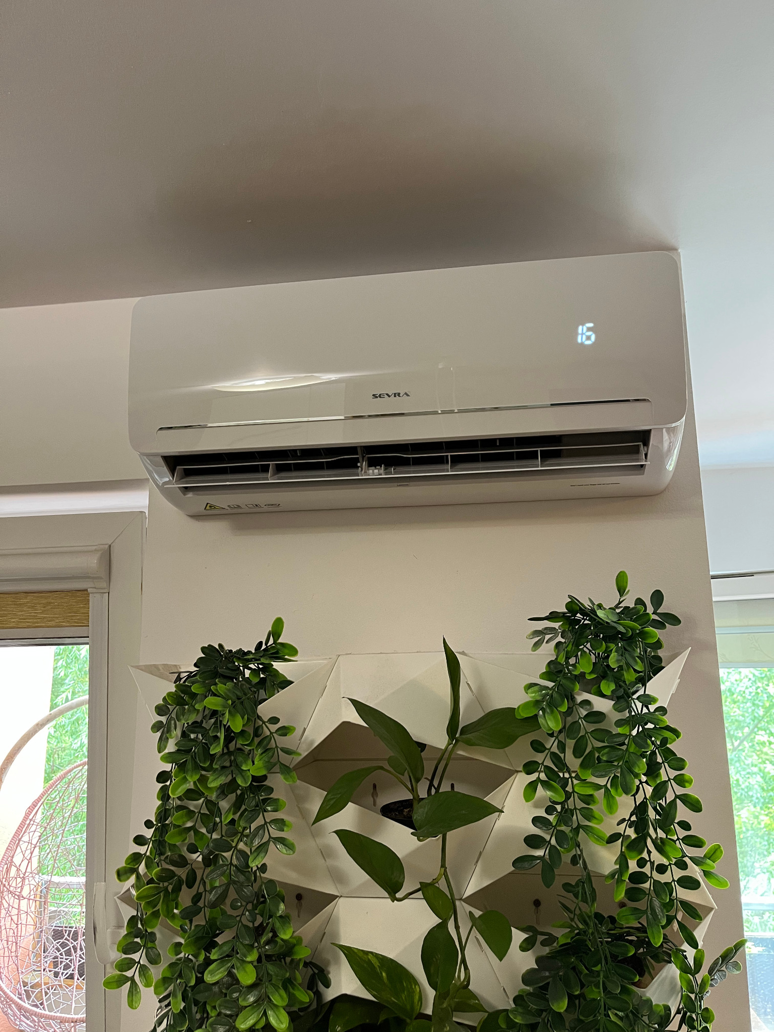 Biały klimatyzator ścienny marki Seyra z wyświetloną temperaturą 16 stopni, umieszczony nad dekoracją z roślin w doniczkach, w jasnym wnętrzu.
