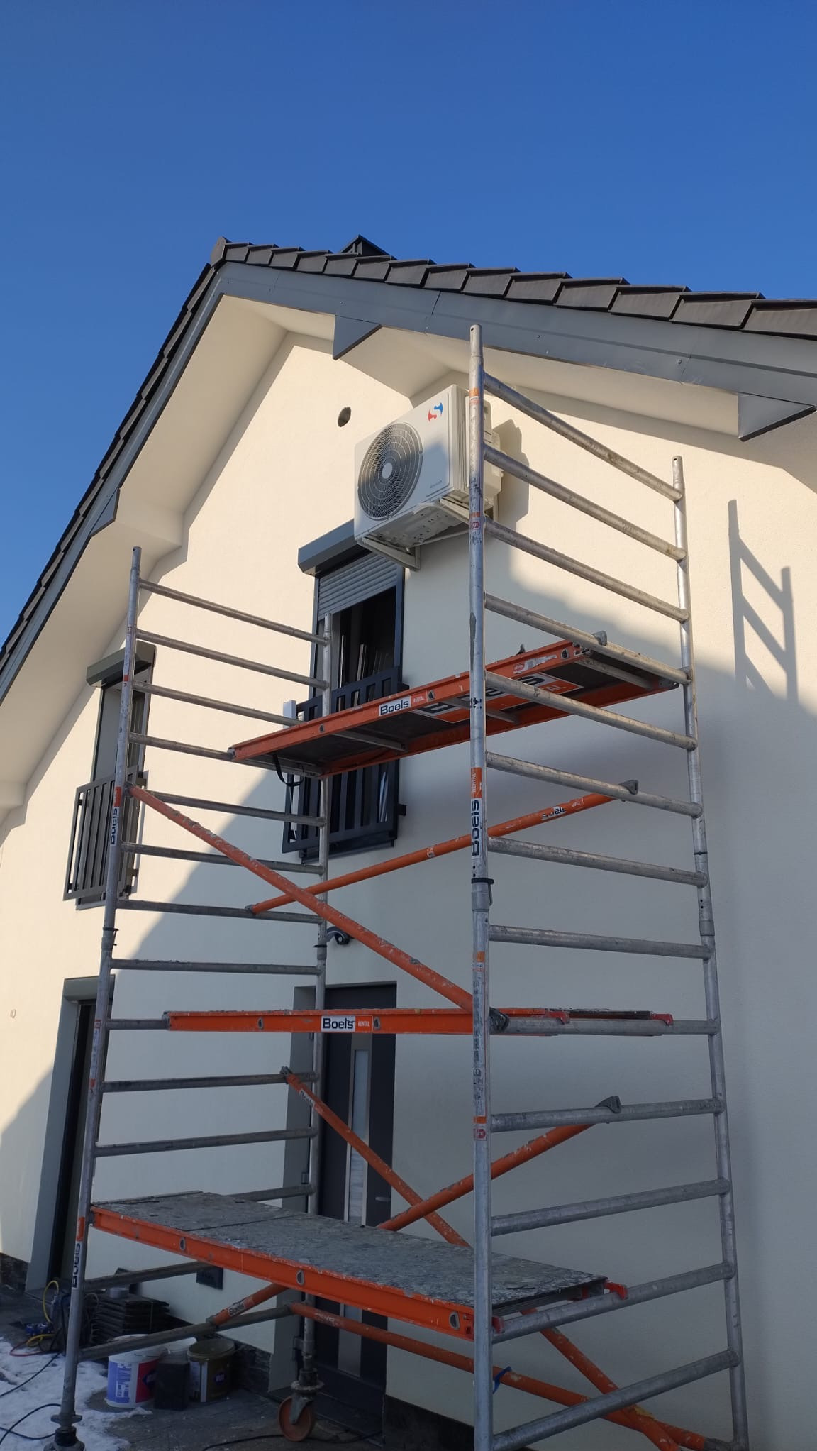 Biały klimatyzator na ścianie domu, zamontowany nad oknem z roletą, widoczny ze rusztowania na tle błękitnego nieba.